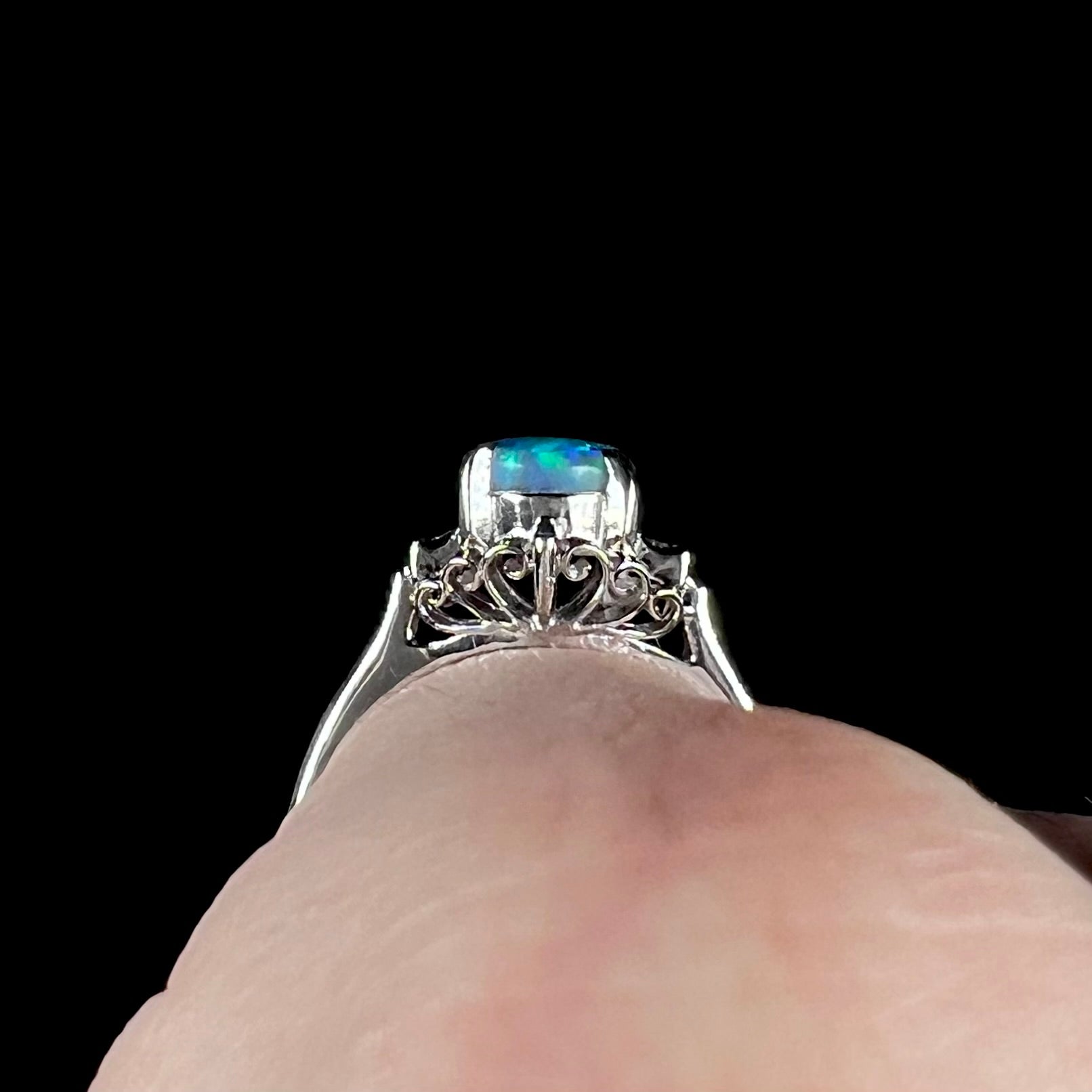 A petite black opal ring worn on a pinkie finger, showcasing the ring's open platinum filigree design.