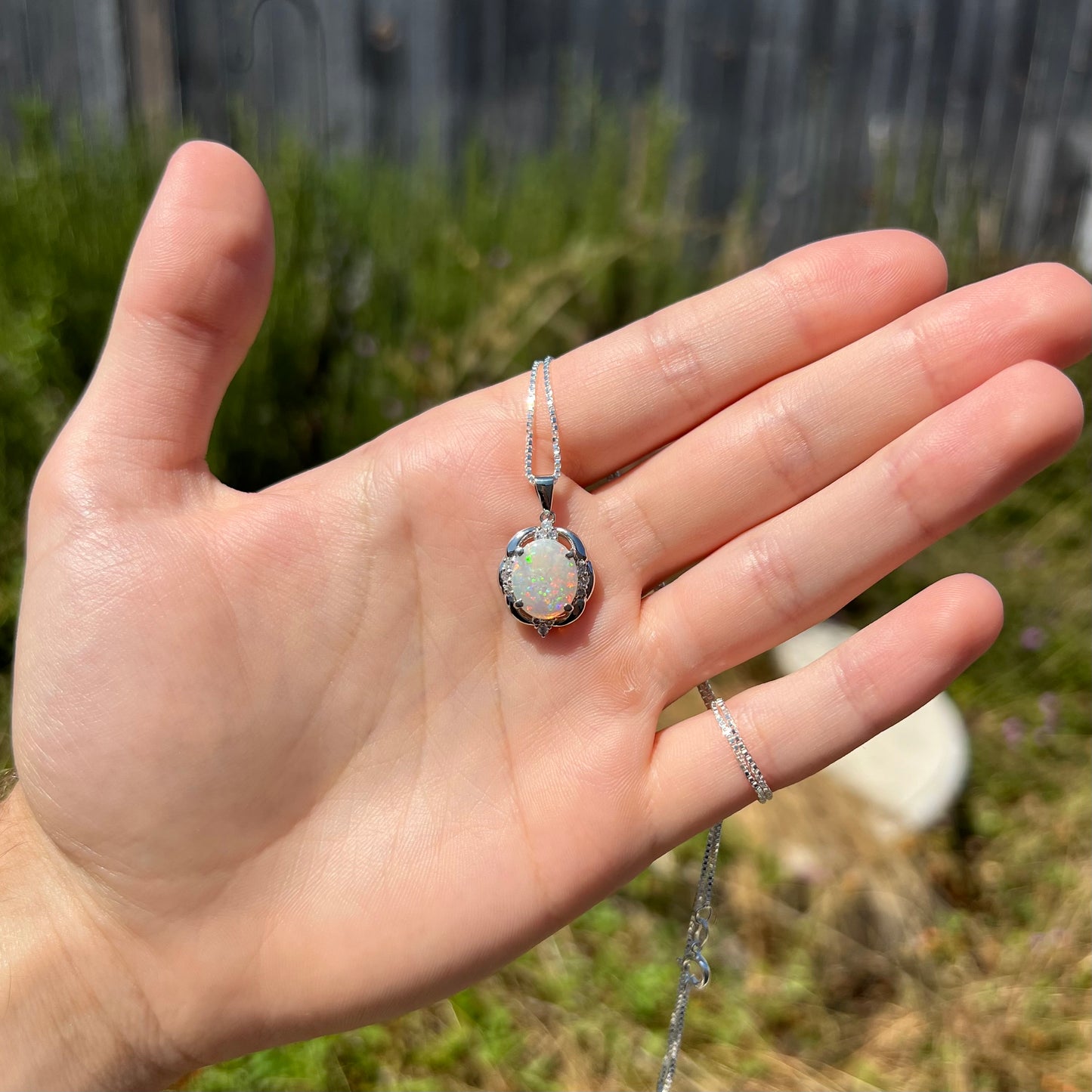 A platinum opal and diamond necklace held in a hand, shown at a distance in the sunlight.