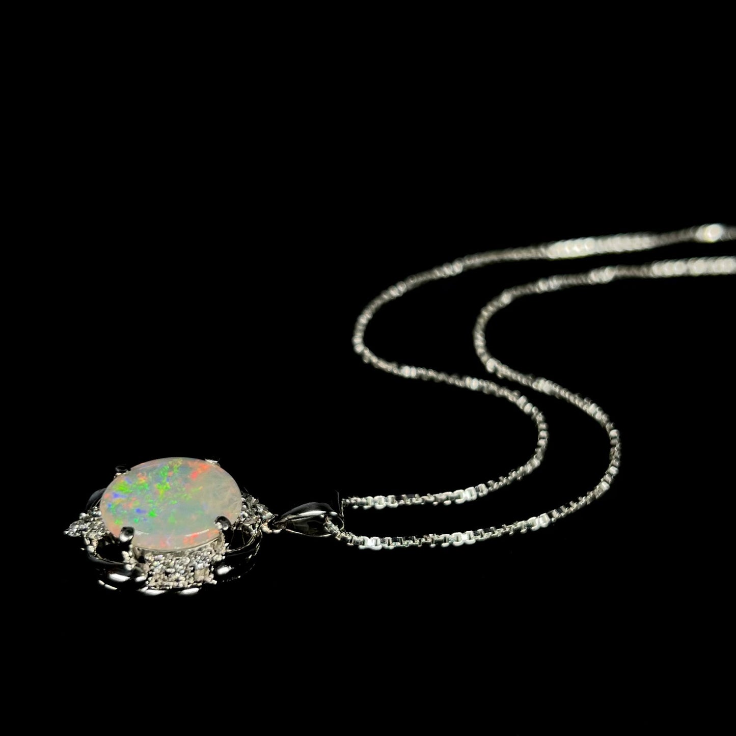 A platinum opal and diamond necklace, laying on a black backdrop, with the chain displayed in a wavy fashion.