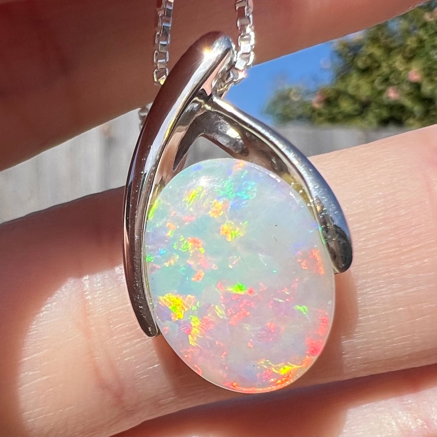 A macro close-up of the platinum opal necklace in sunlight, tilted slightly, with dominant red and yellow fire against a finger.