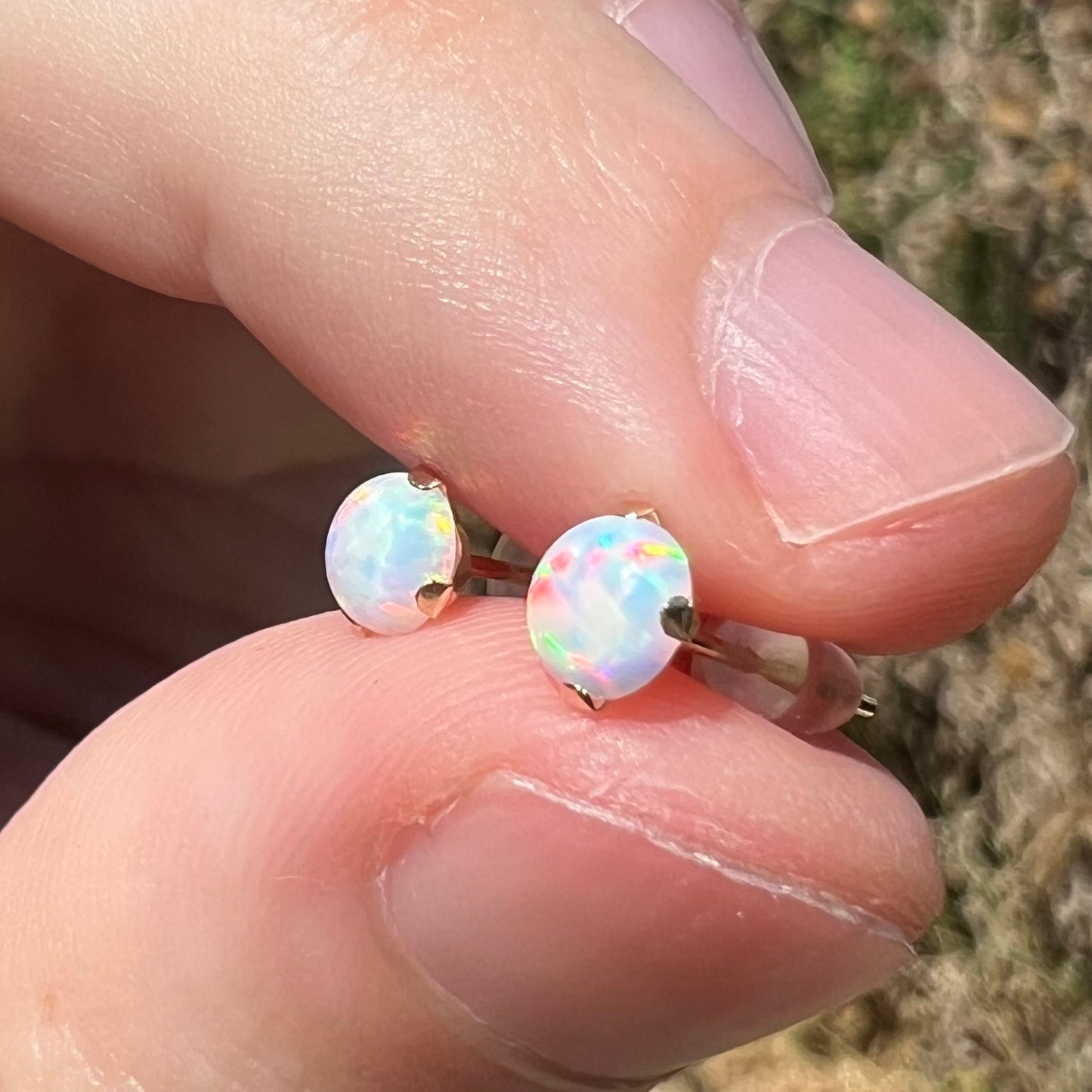 A pair of prong-set pushback lab-created opal stud earrings in 14k gold, held between two fingers and shown close-up from an angled view in sunlight.