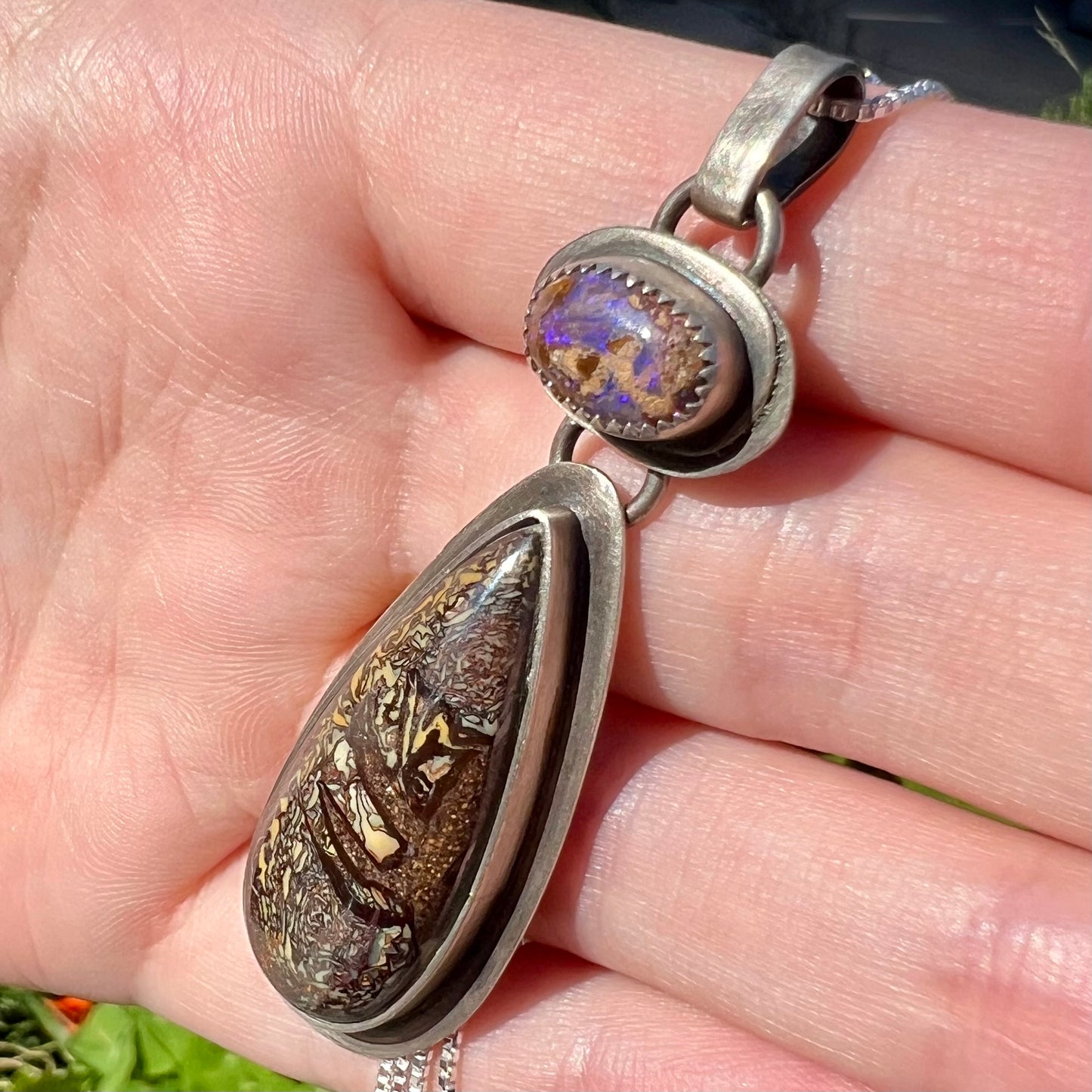 Sterling silver two-stone Australian boulder opal pendant held in a hand in sunlight. The top opal has purple coloring in the light.