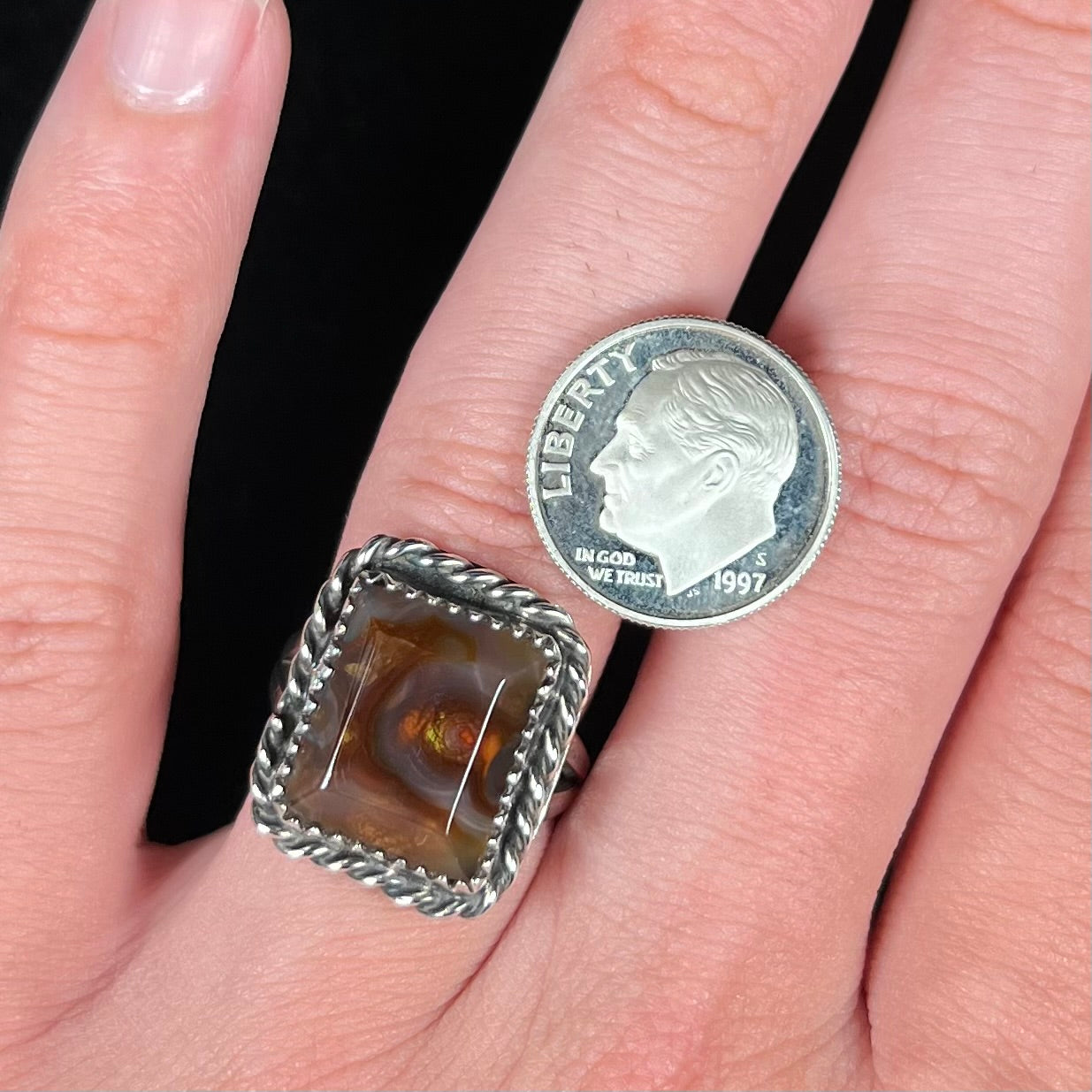 Close-up of a rectangle shaped fire agate ring worn on a finger next to a dime coin for scale.