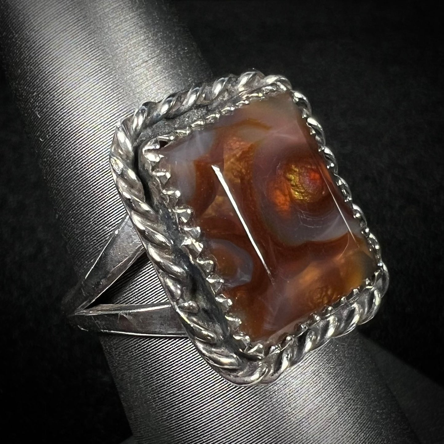 Angled view of sterling silver rectangle shaped fire agate ring with rope design against dark background.