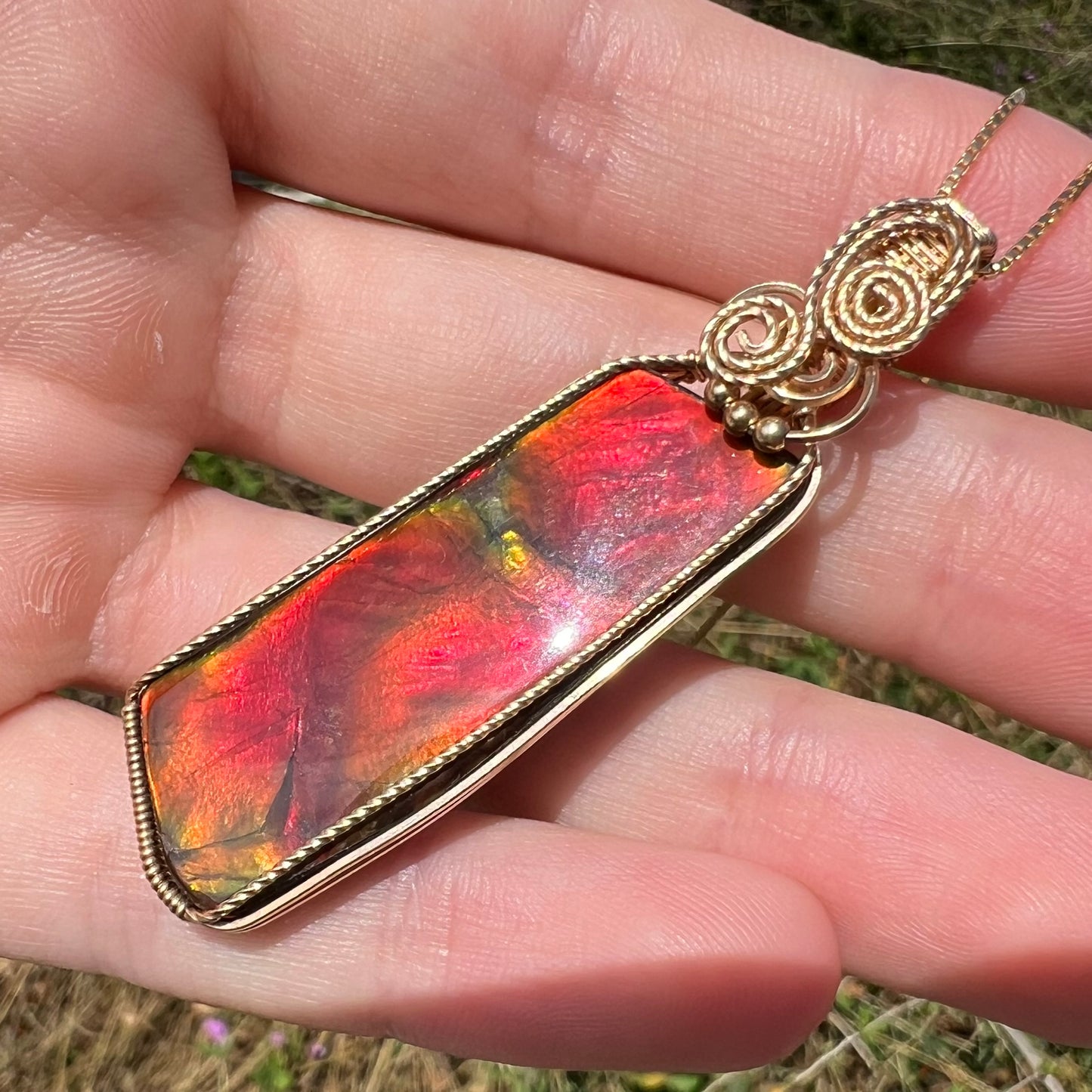 A red ammolite pendant with a gold filled wire wrapped design held at an angle in the sunlight by a hand.