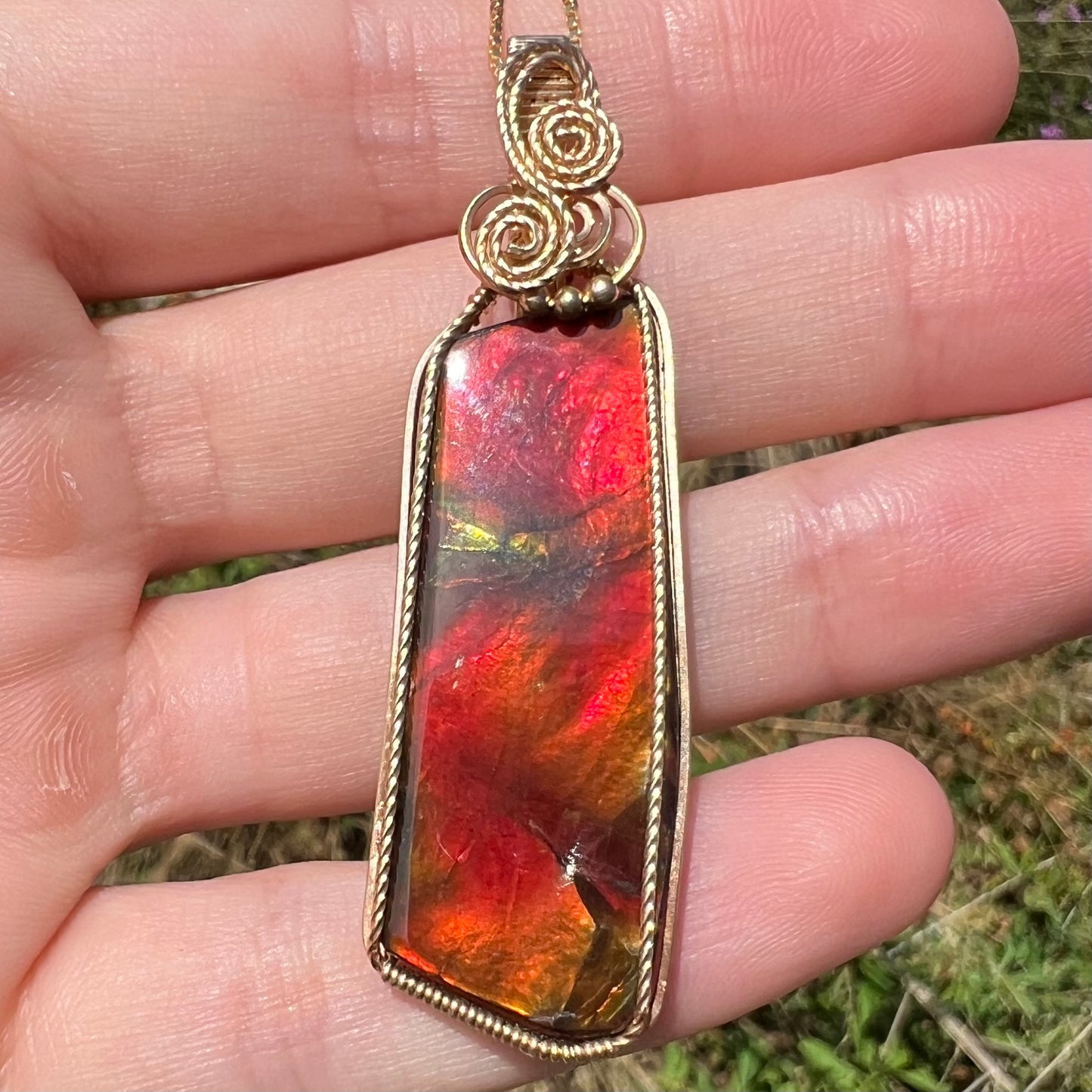 Front straight close-up view of a red ammolite pendant held in the sunlight by a hand.