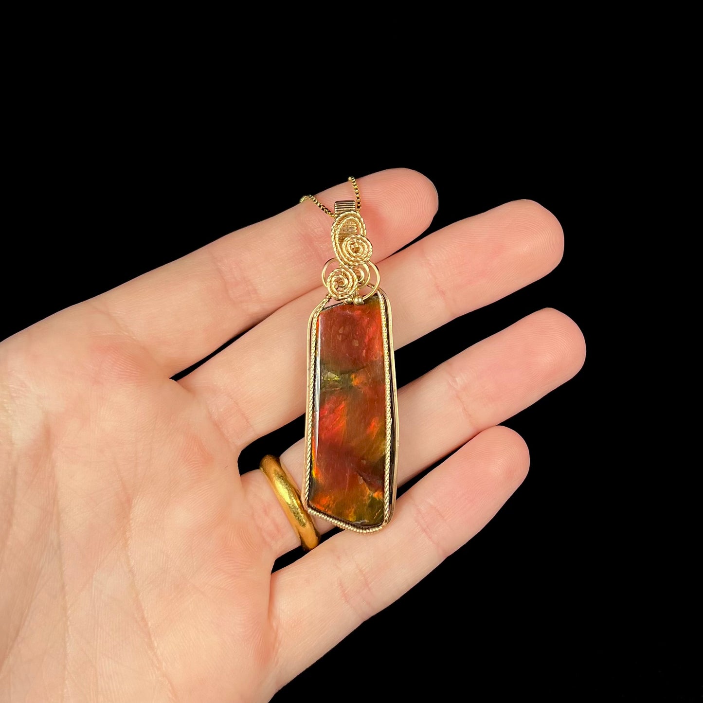 A red ammolite wire wrapped pendant held in a hand against a black background.