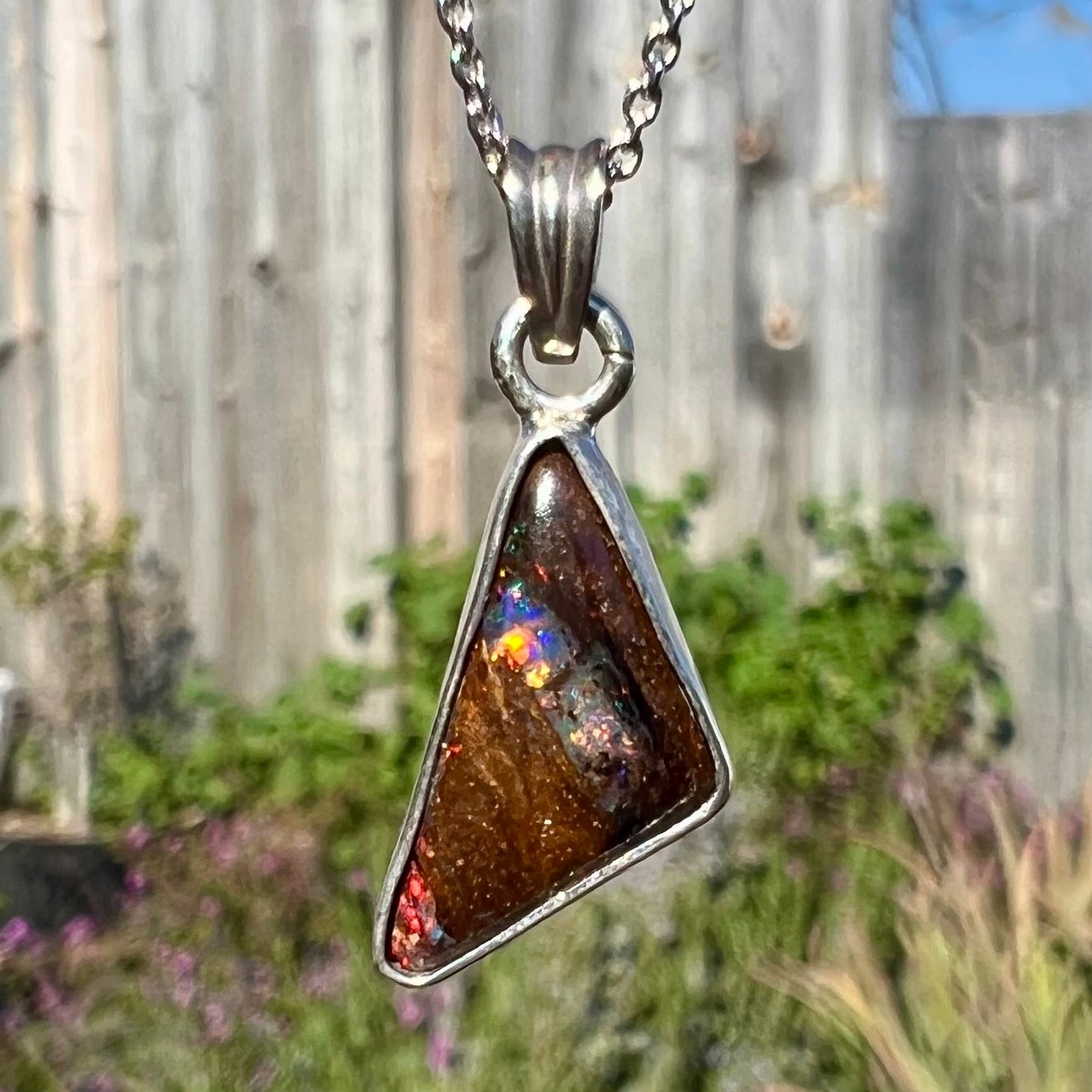 A sterling silver necklace bezel set with a triangle shaped boulder opal.  The opal has red and blue flashes.