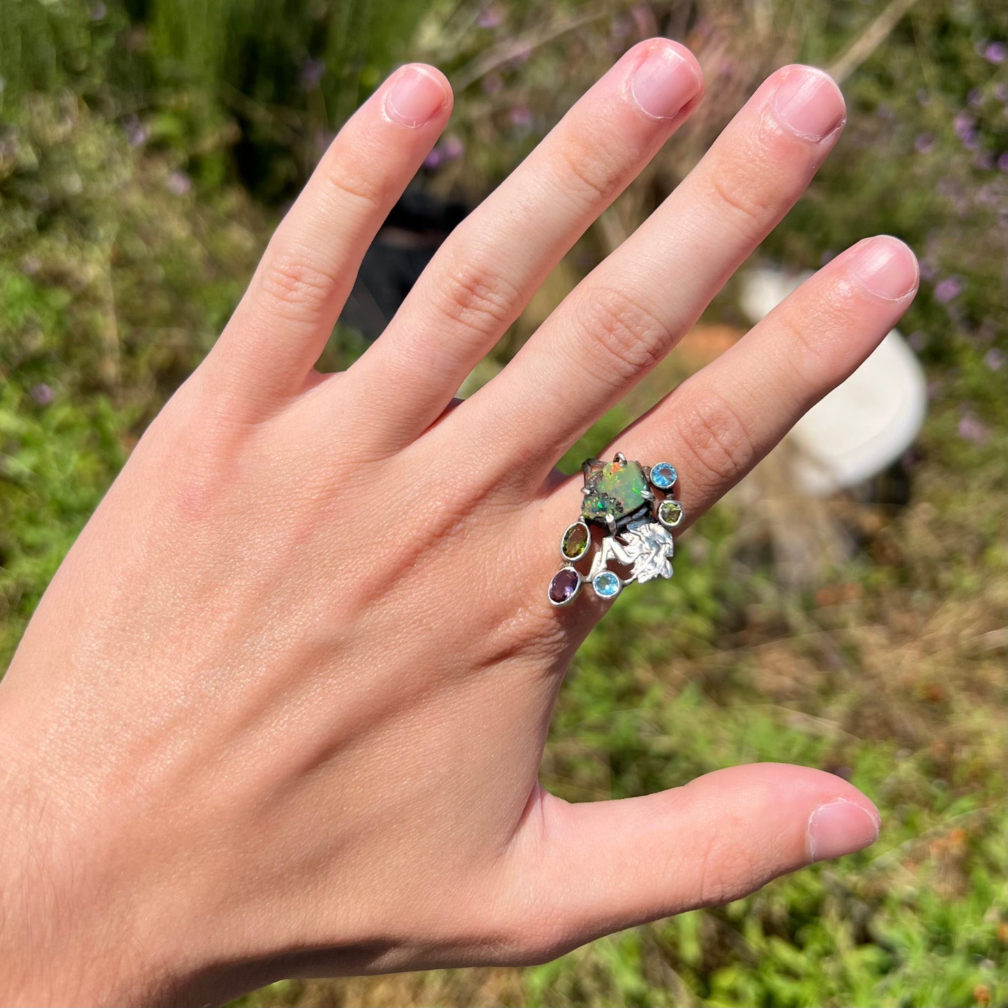A silver men's ring set with rough Virgin Valley opal and gems with a female figure design worn on a hand in sunlight.