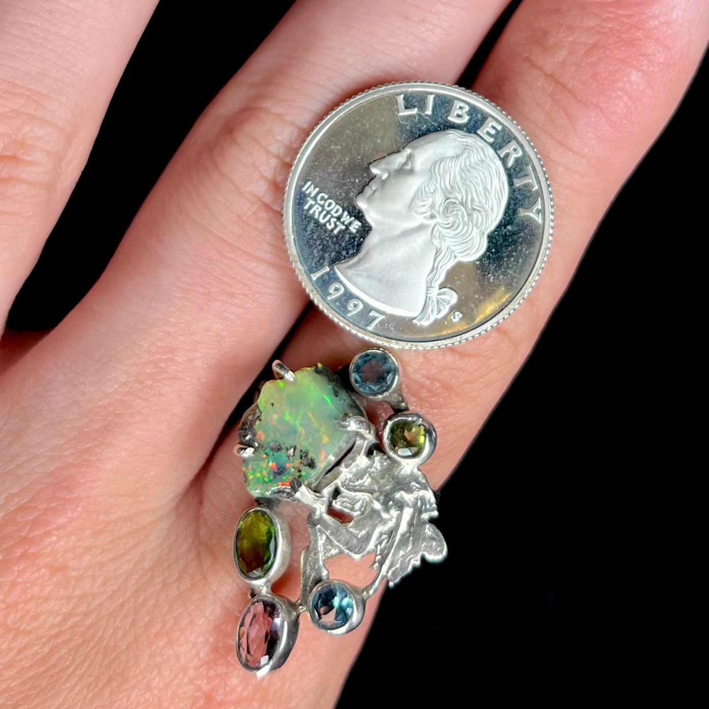 A silver men's ring set with rough Virgin Valley opal and gems with a female figure design worn next to a quarter coin.