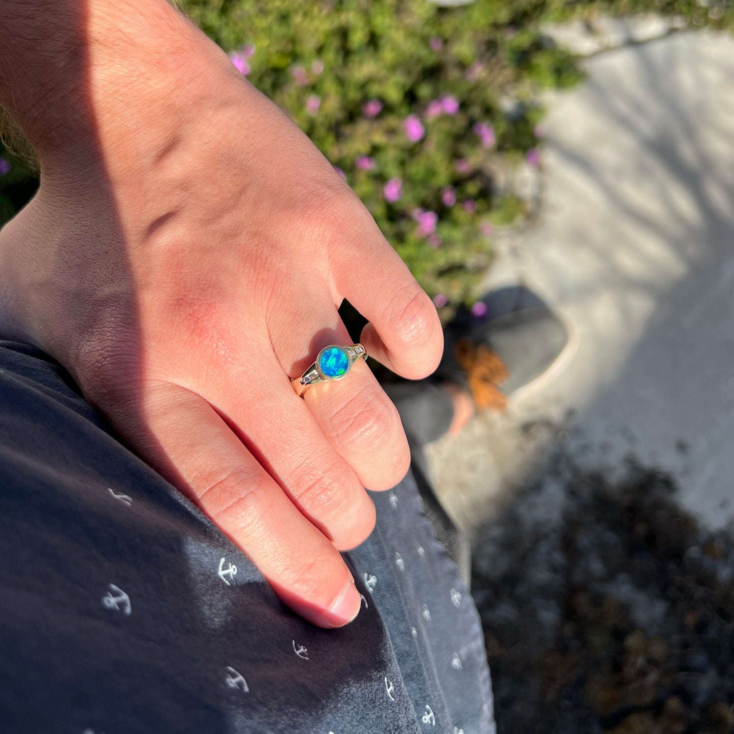 A hand resting on a model's hip while wearing a round boulder opal ring with a blue body tone. The opal shines with green color play in the sunlight.