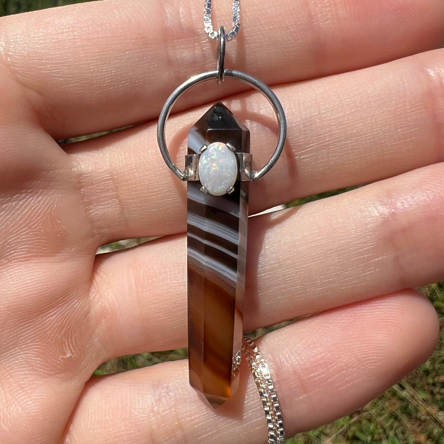 A sardonyx and opal pendant shown in a hand from the front close-up in the sunlight.