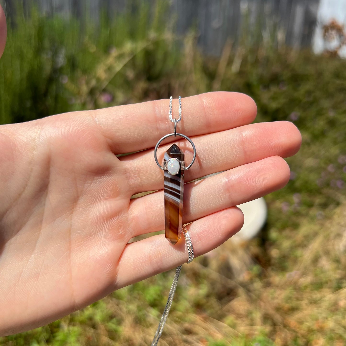 A sardonyx and opal pendant made from silver, held in the sunlight by a hand.