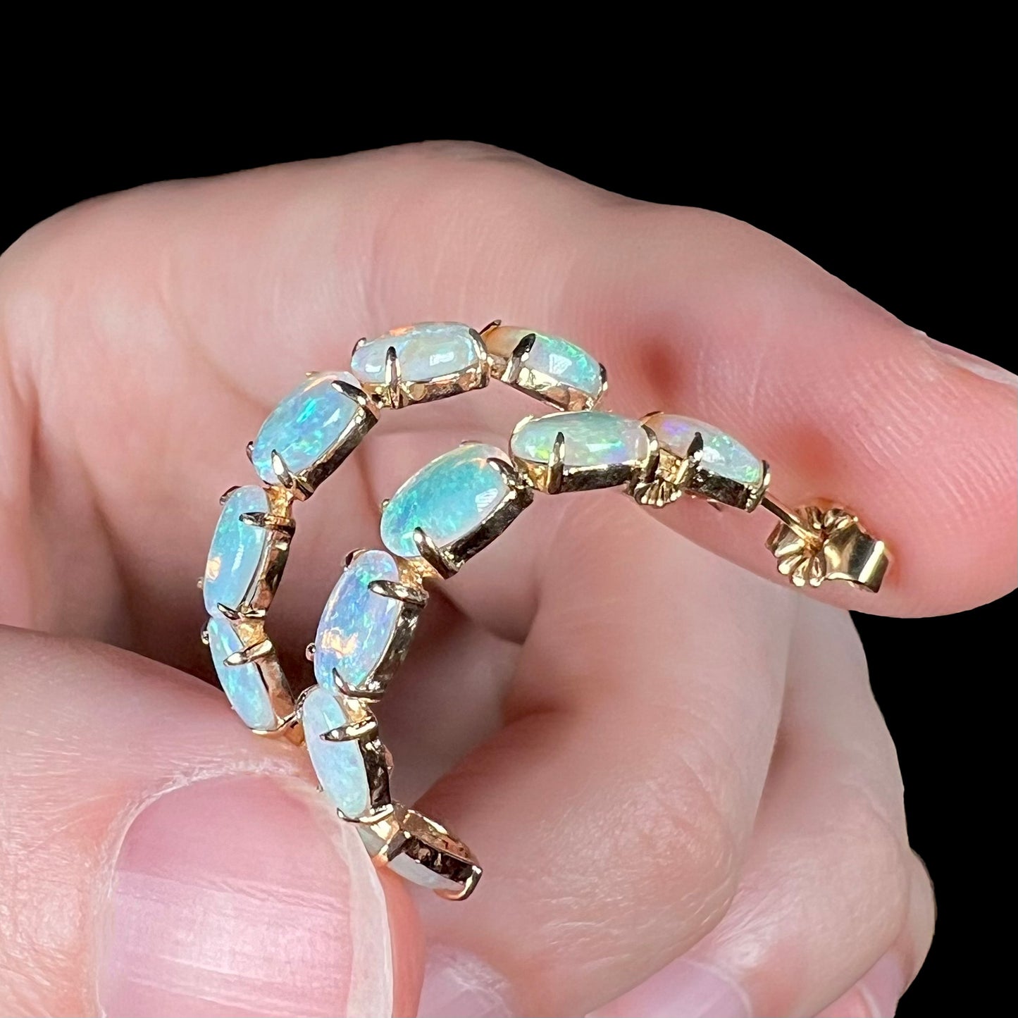 Opposite angle of semi-hoop opal earrings held in fingers, green and orange play visible.
