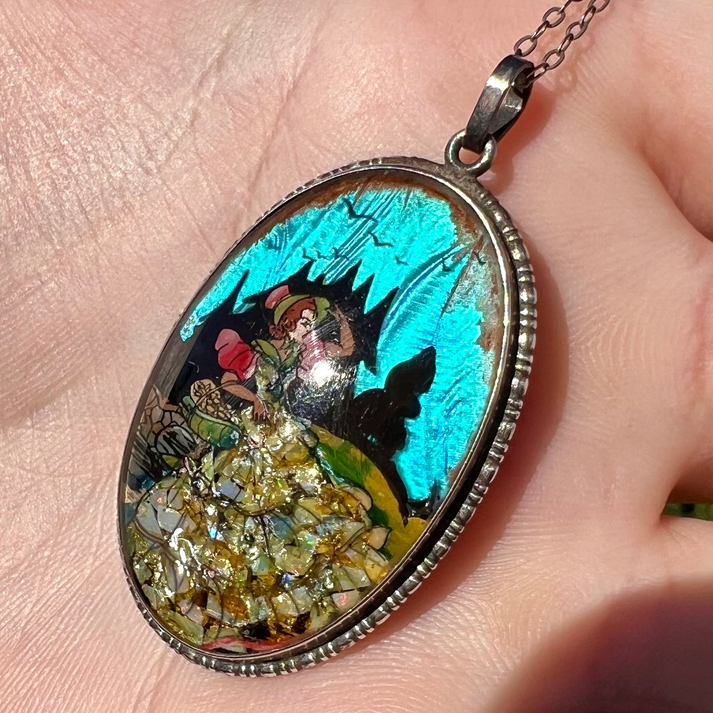 A silver pendant with blue butterfly wing and crushed opal, shown close-up in a hand at an angle in sunlight.