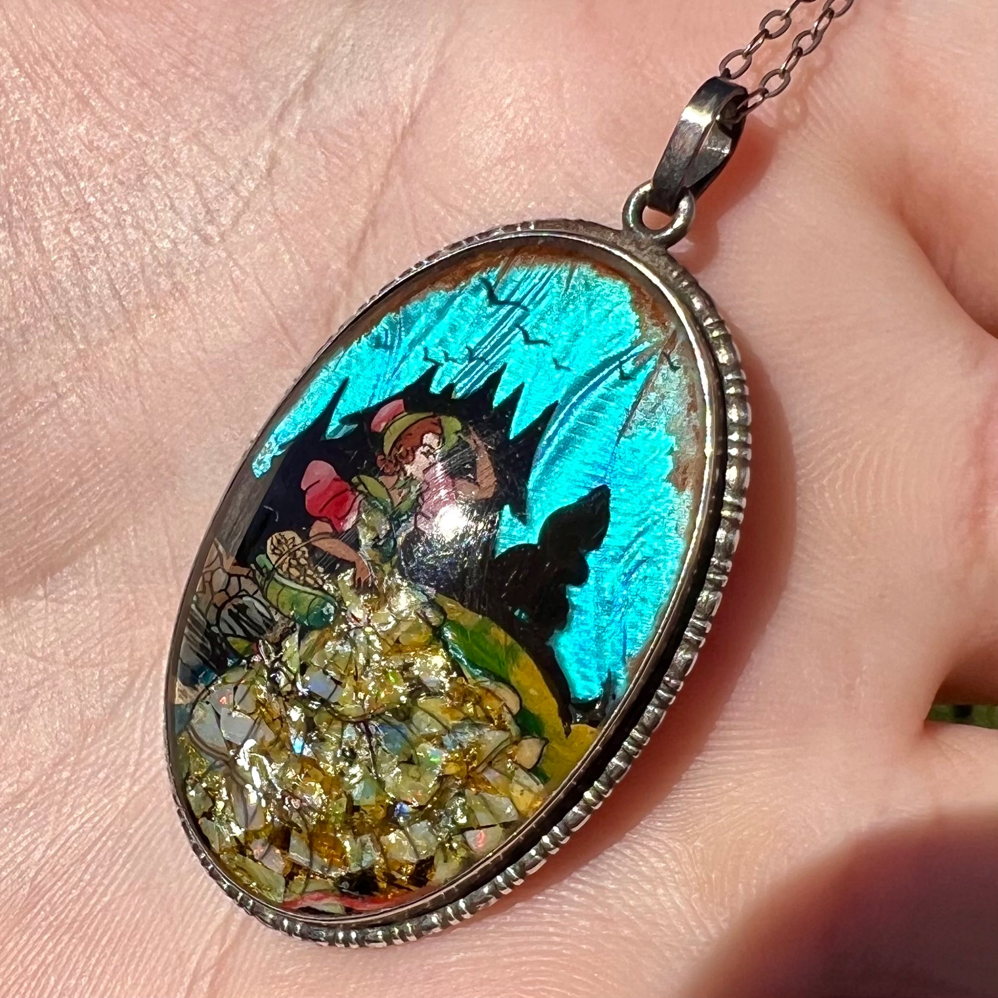 A silver pendant with blue butterfly wing and crushed opal, shown close-up in a hand at an angle in sunlight.