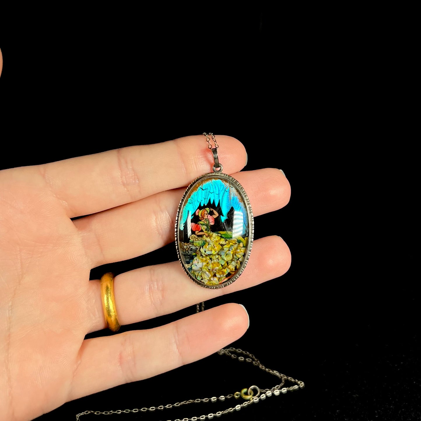 A sterling silver butterfly wing and opal necklace with the picture of a woman, held in a hand against black background.