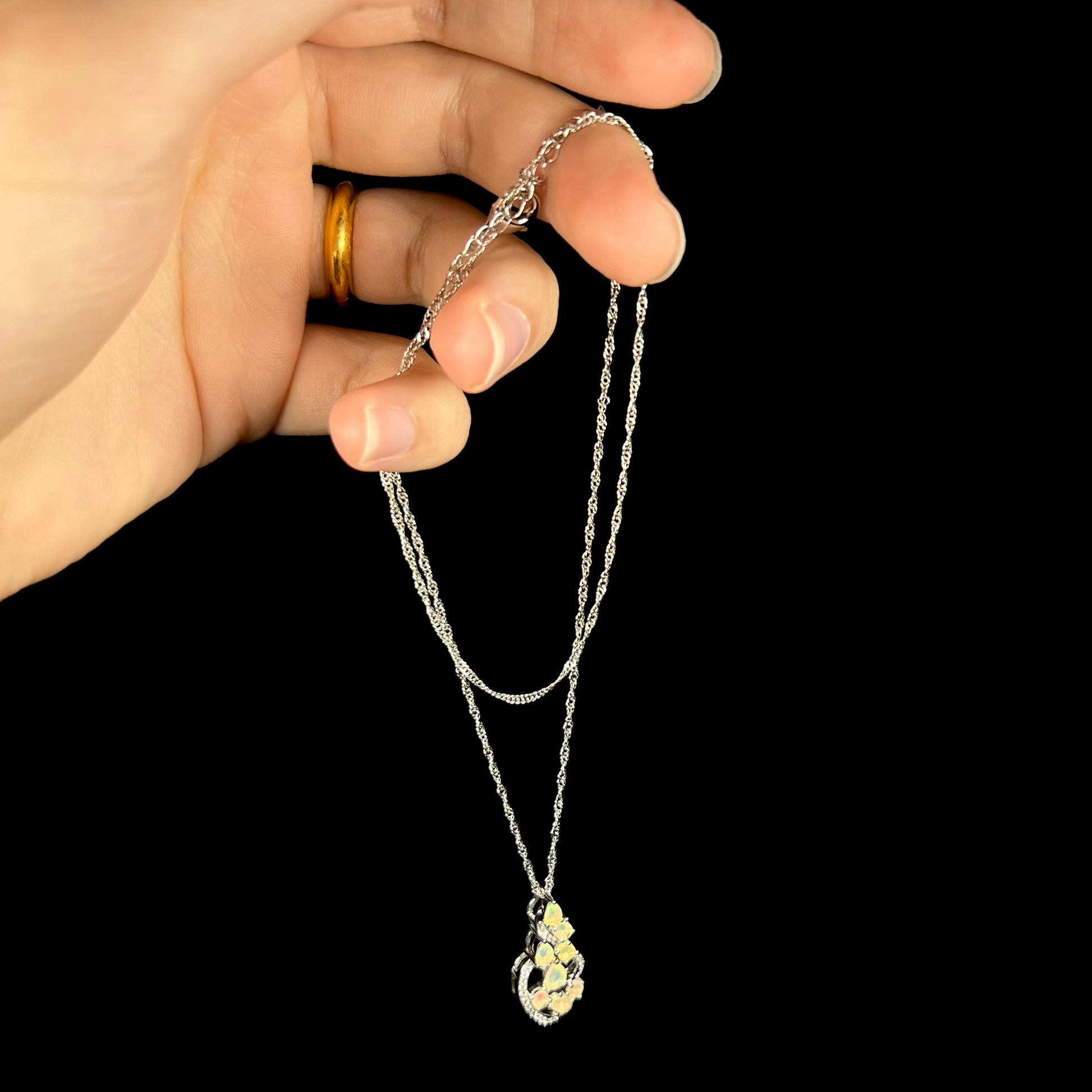 A silver Ethiopian opal necklace dangling from a hand by its chain against a black backdrop.