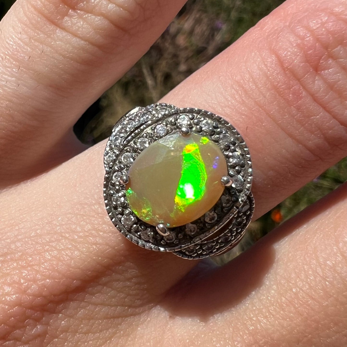 Close-up front view of a silver Ethiopian opal ring worn on a finger in sunlight. The opal has a green flash.