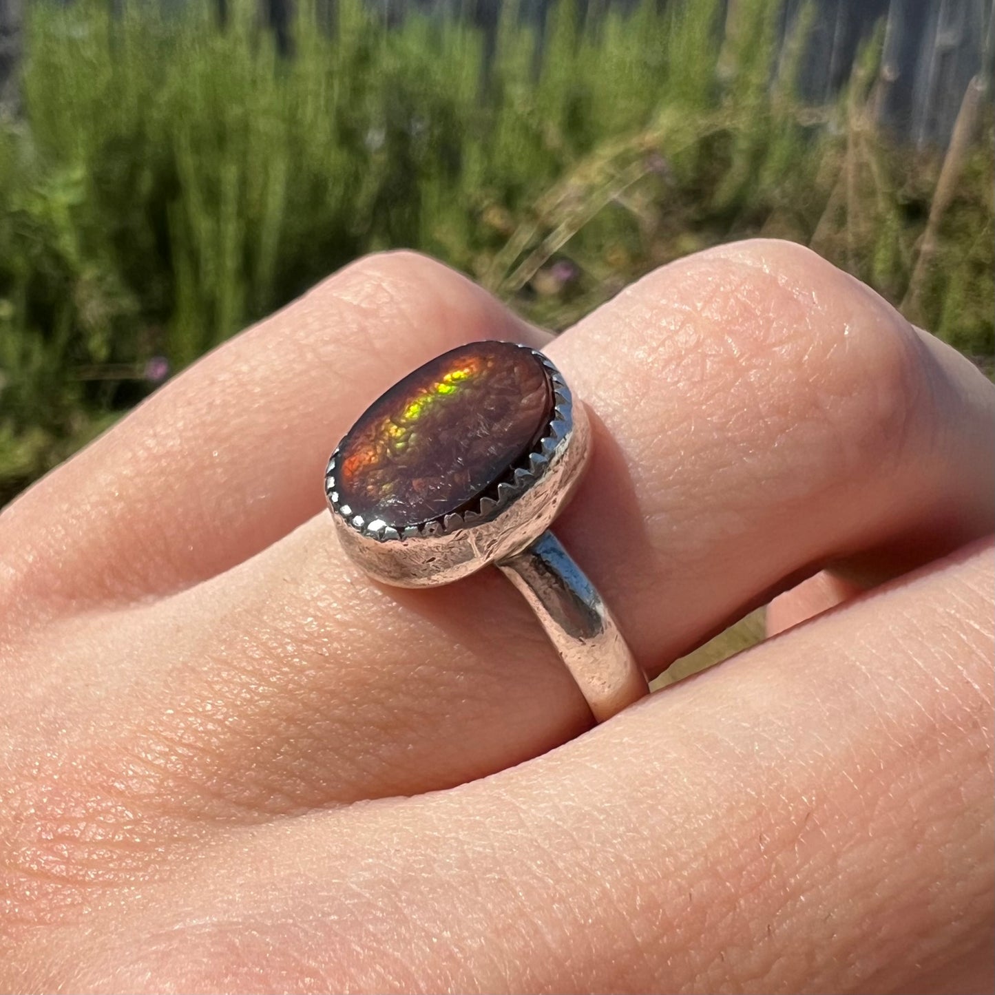 A silver fire agate ladies' ring worn on a finger in the outdoor sunlight, shown at an angled view.
