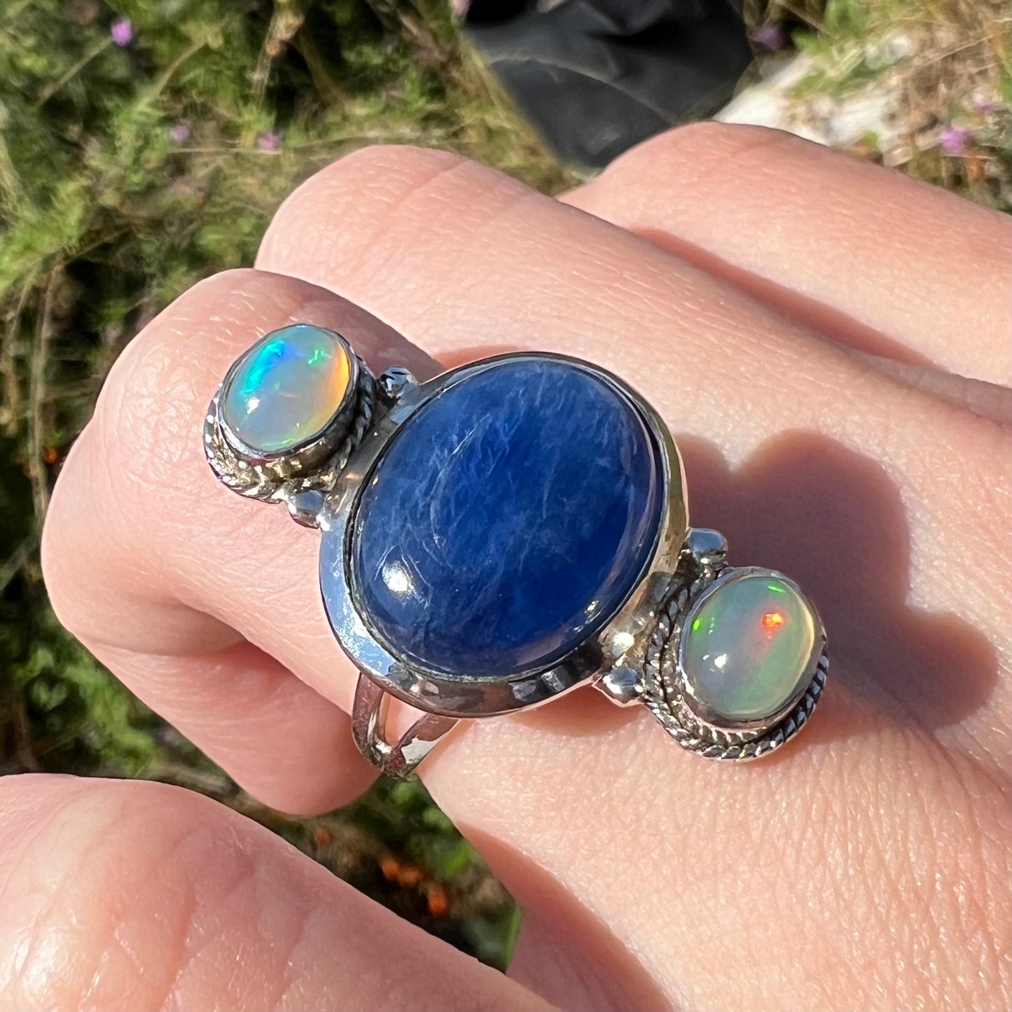 Macro view of a silver kyanite and opal ring worn on a finger in the sunlight, shown at an angle.
