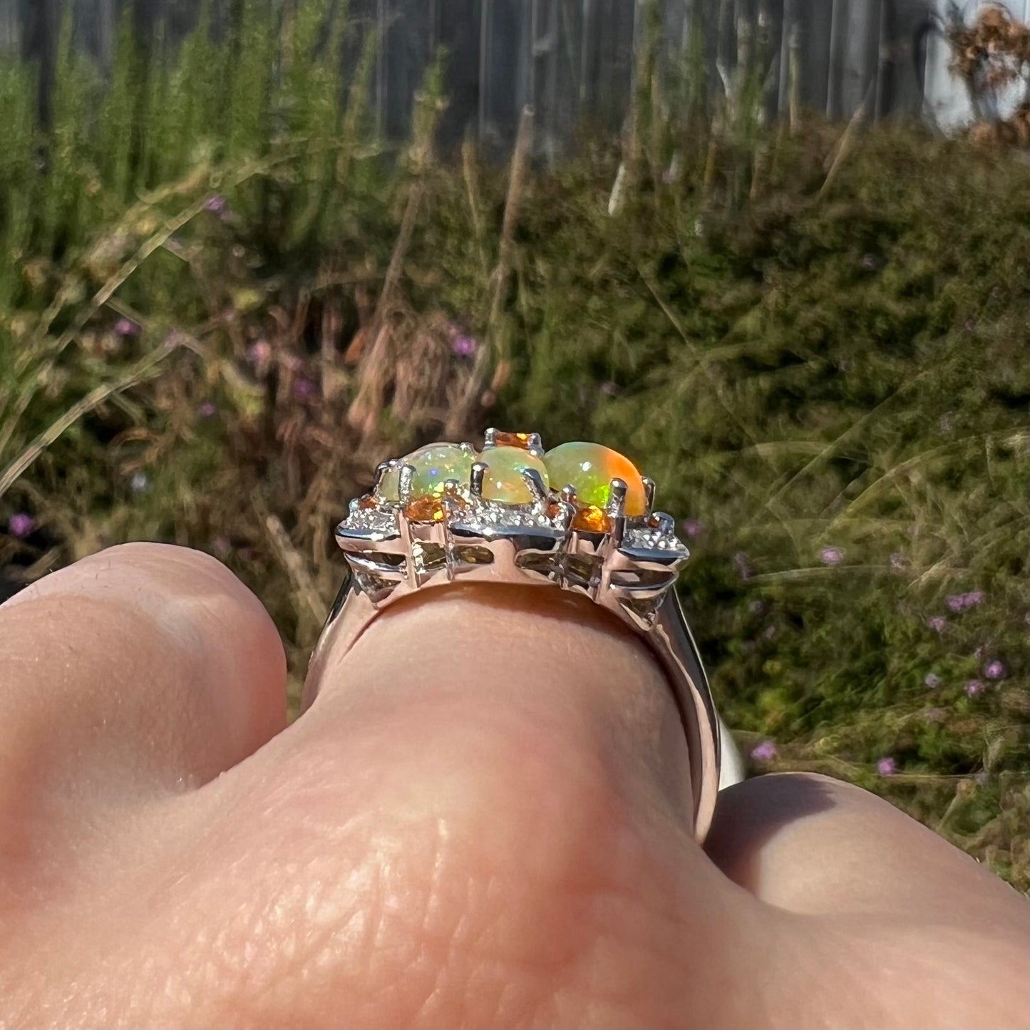 Side gallery profile view of a silver opal cluster ring in natural light, worn on a finger against a blurred green backdrop.