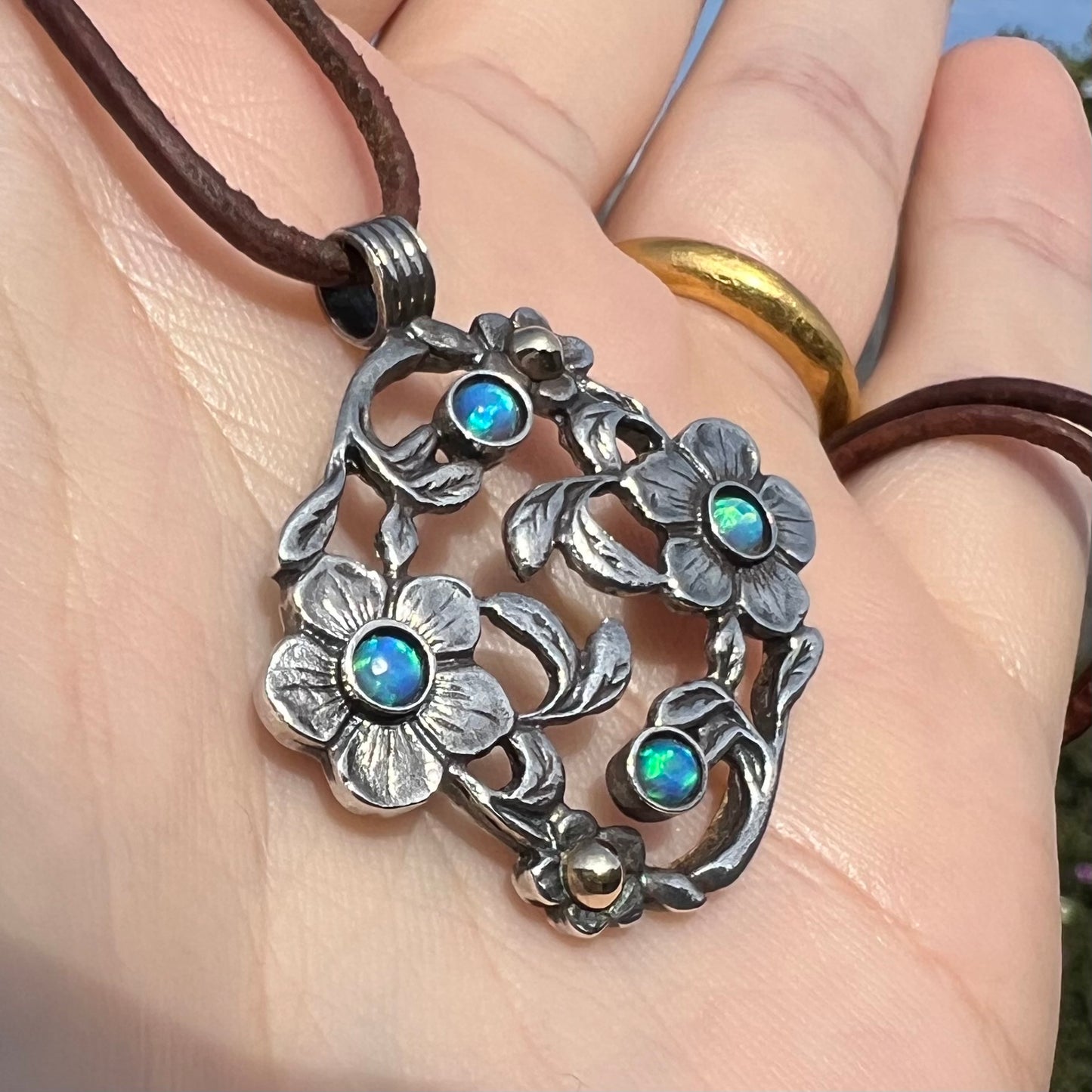 A two-tone sterling silver and 14k gold opal dogwood flower pendant shown from an angle in the sunlight held in a hand.