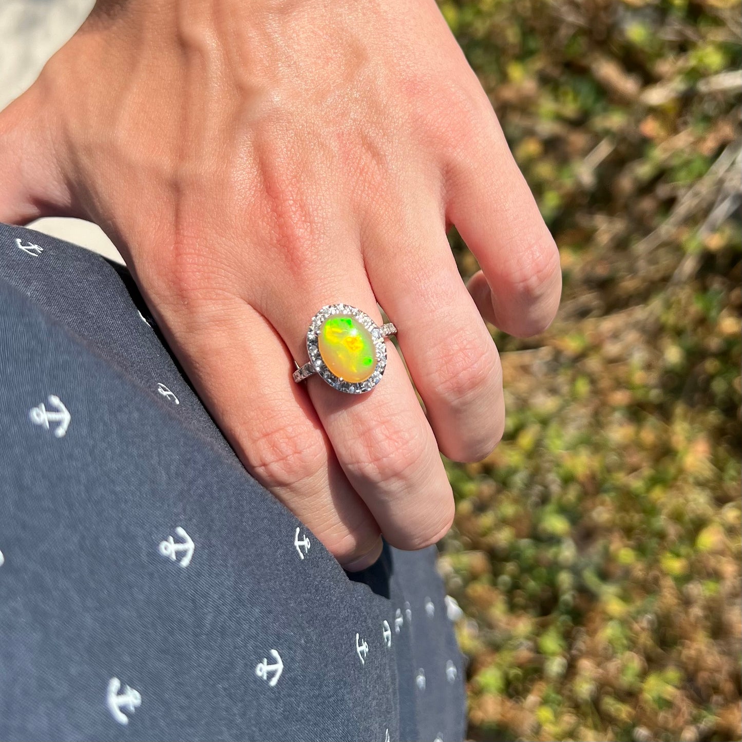 An Ethiopian fire opal ring with a CZ halo and green color play worn on a hand, resting on the model's hip.