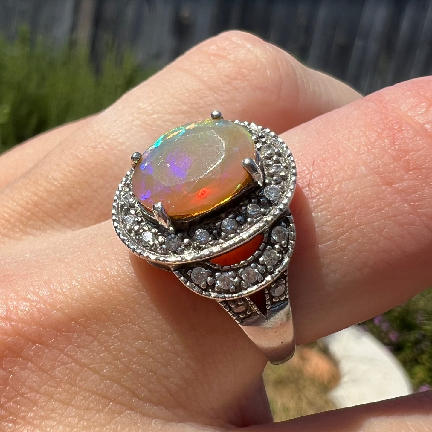 Angled macro view of a silver opal halo-style ring, worn on a finger in natural light.