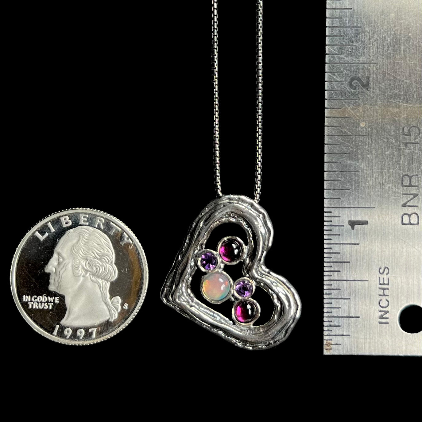 A silver heart necklace set with opal, amethysts, and rhodolite garnets, shown next to a ruler and quarter.