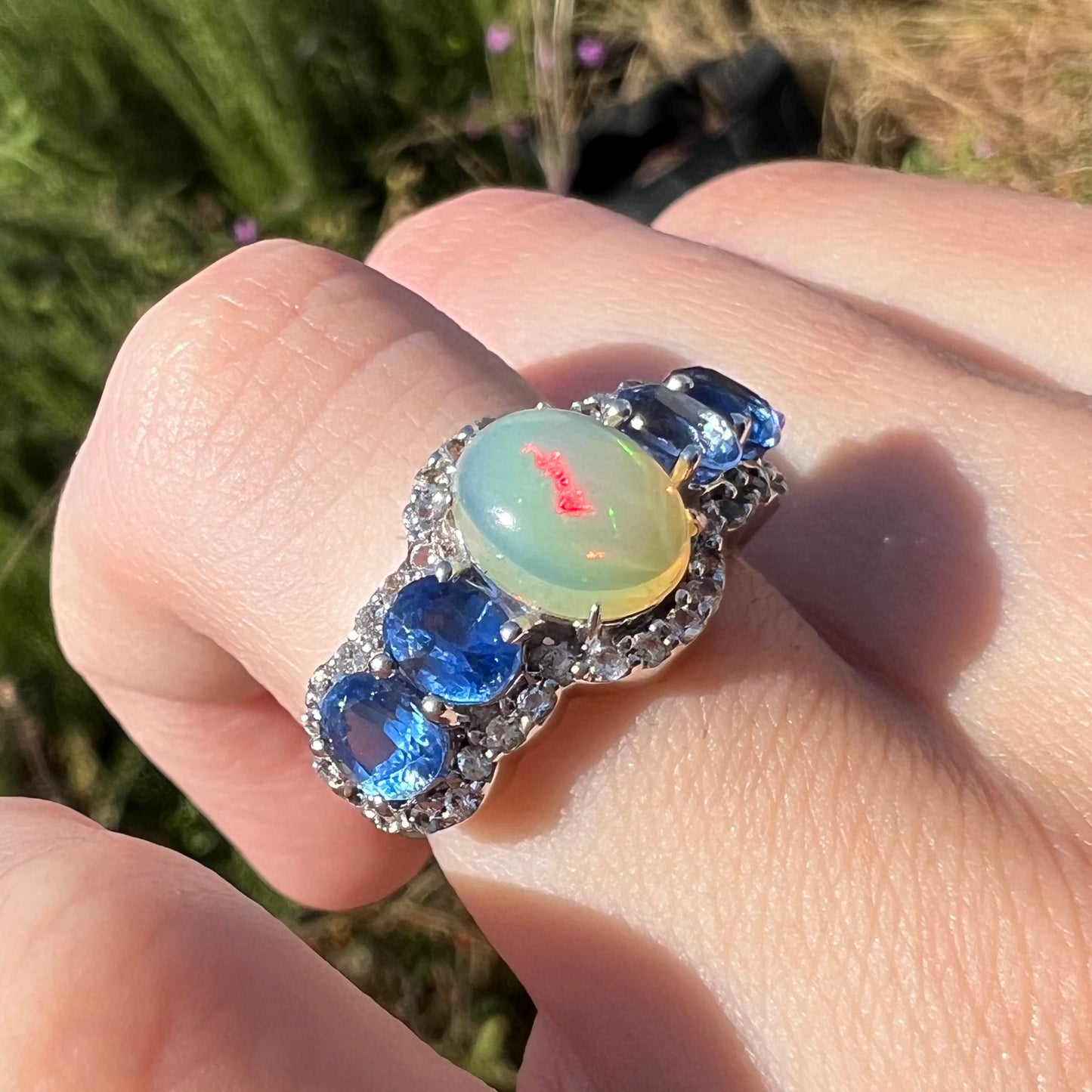 Close-up of a silver Ethiopian opal and kyanite ring worn on a hand in the sunlight. The opal has a red flash.