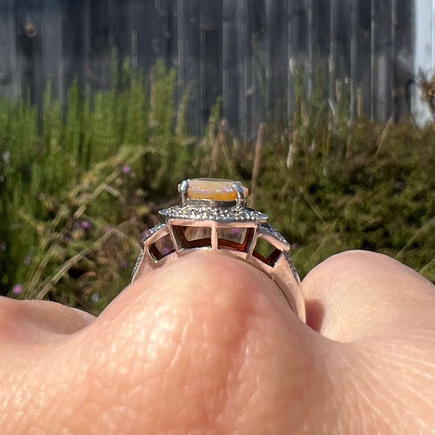 Side gallery view of silver opal ring worn on a finger in daylight against blurred outdoor background.