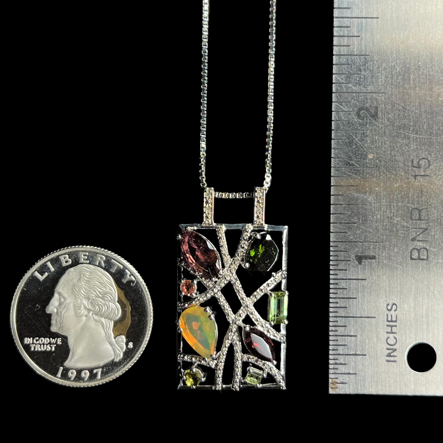 A silver multi-stone necklace set with opal, tourmaline, and diamonds, shown next to a quarter coin and ruler.