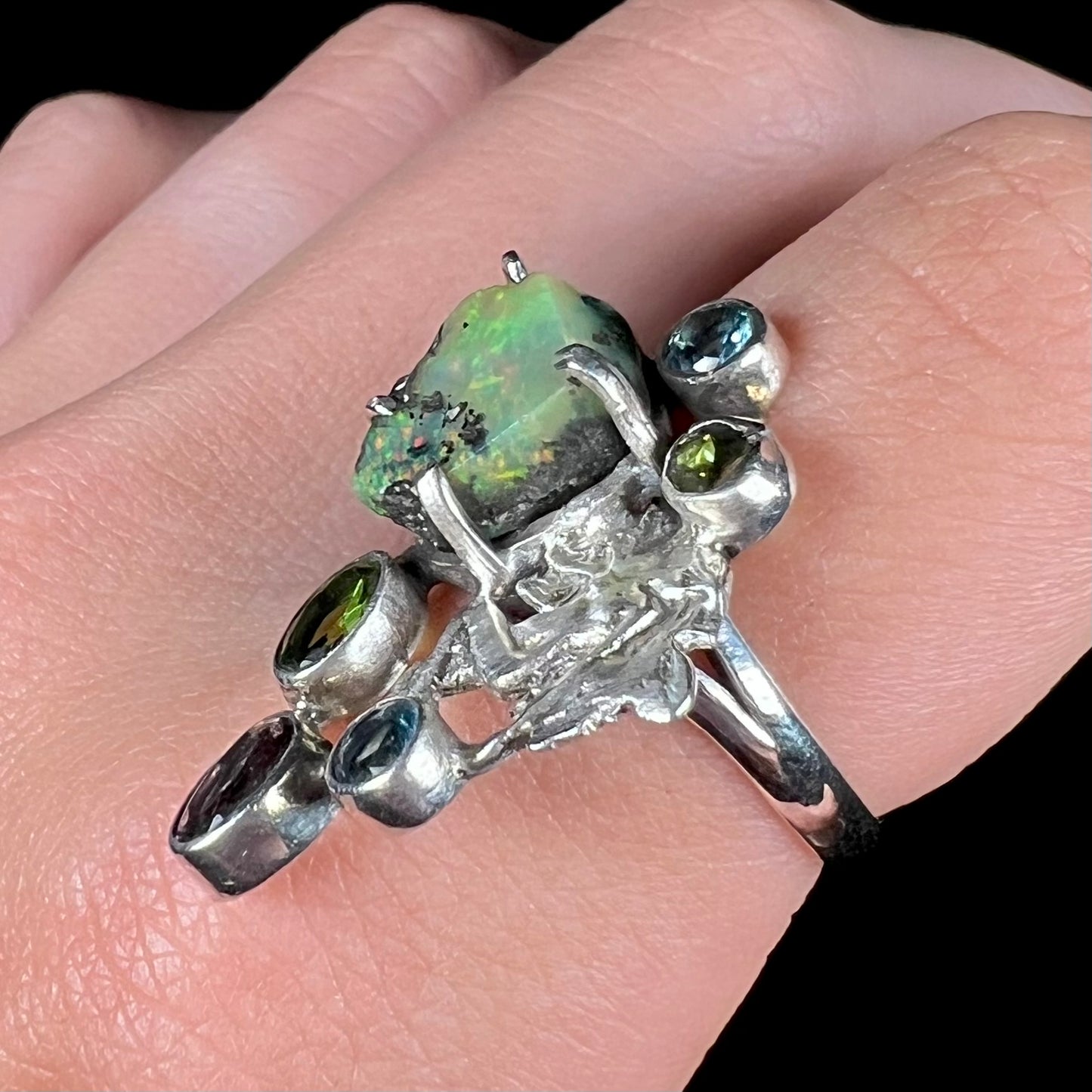 Angled view of a silver men's ring set with rough Virgin Valley opal and gems with a female figure design on a finger.