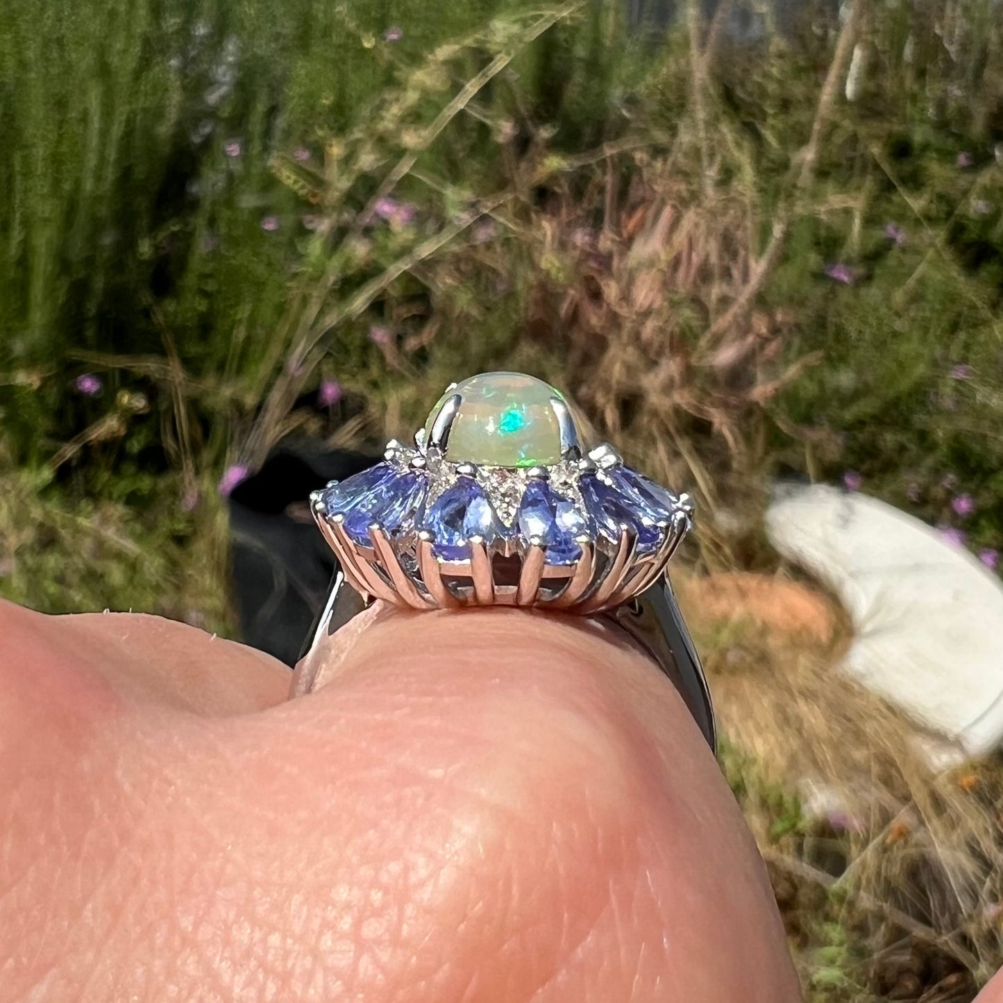 Side gallery view of a silver tanzanite and opal ring, worn on a finger in sun.