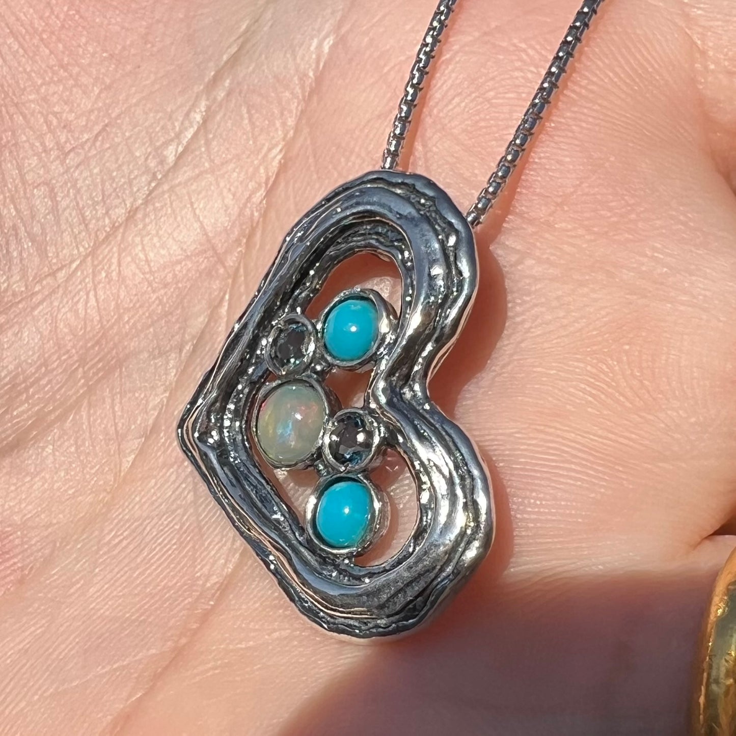 Angled close-up view of a sterling silver heart necklace set with opal, turquoise, and blue topaz shown in sunlight.