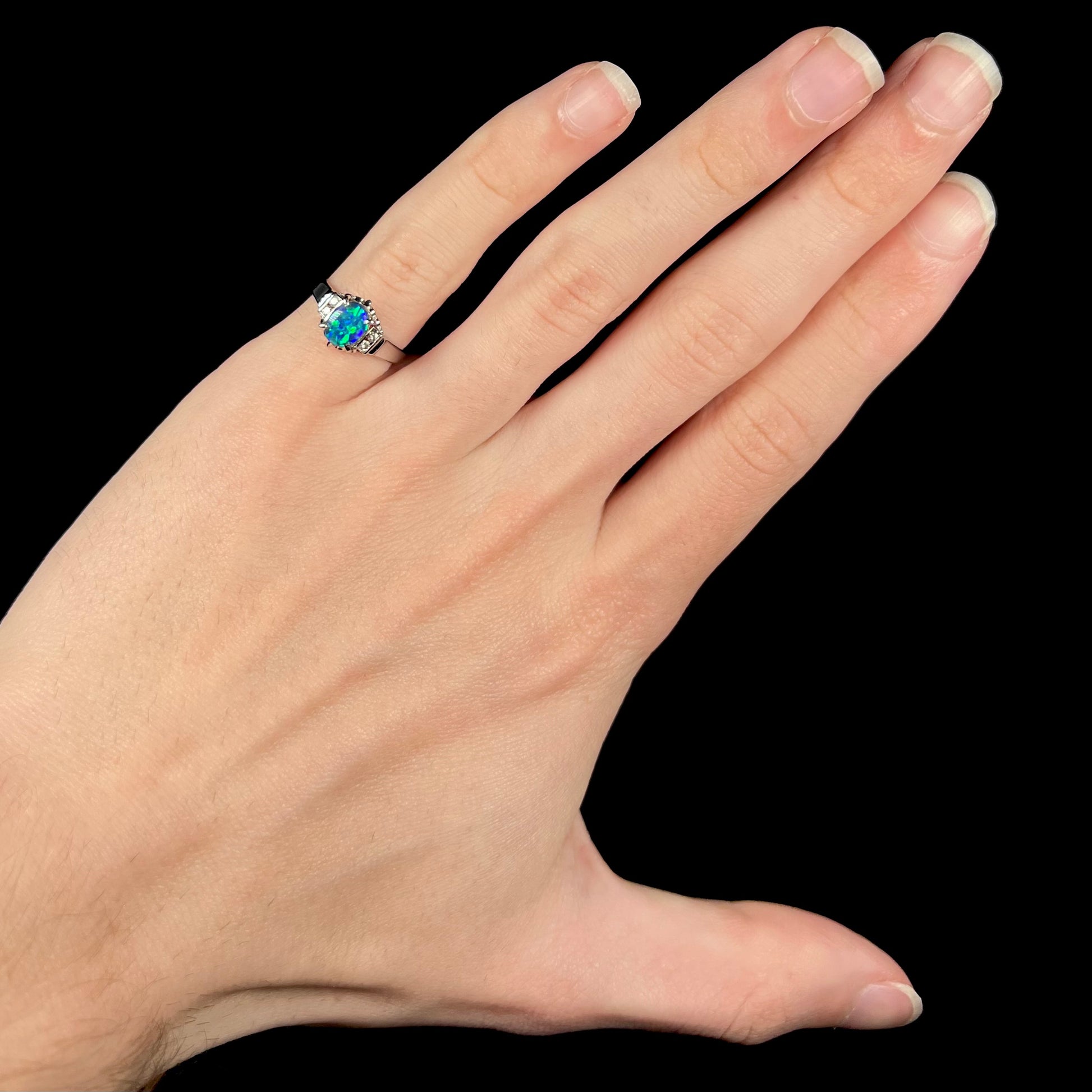 A petite, size 4.5 black opal engagement ring is worn on a hand's pinkie finger. The ring is platinum and has diamond accents.