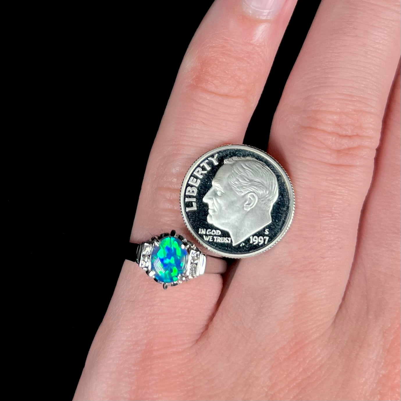 A small black opal platinum ring with vibrant green color play is worn on a pinkie finger next to a US dime coin.
