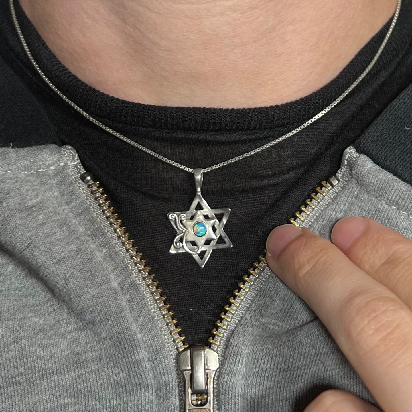 A sterling silver Star of David opal necklace worn against a dark shirt with a partially unzipped jacket.