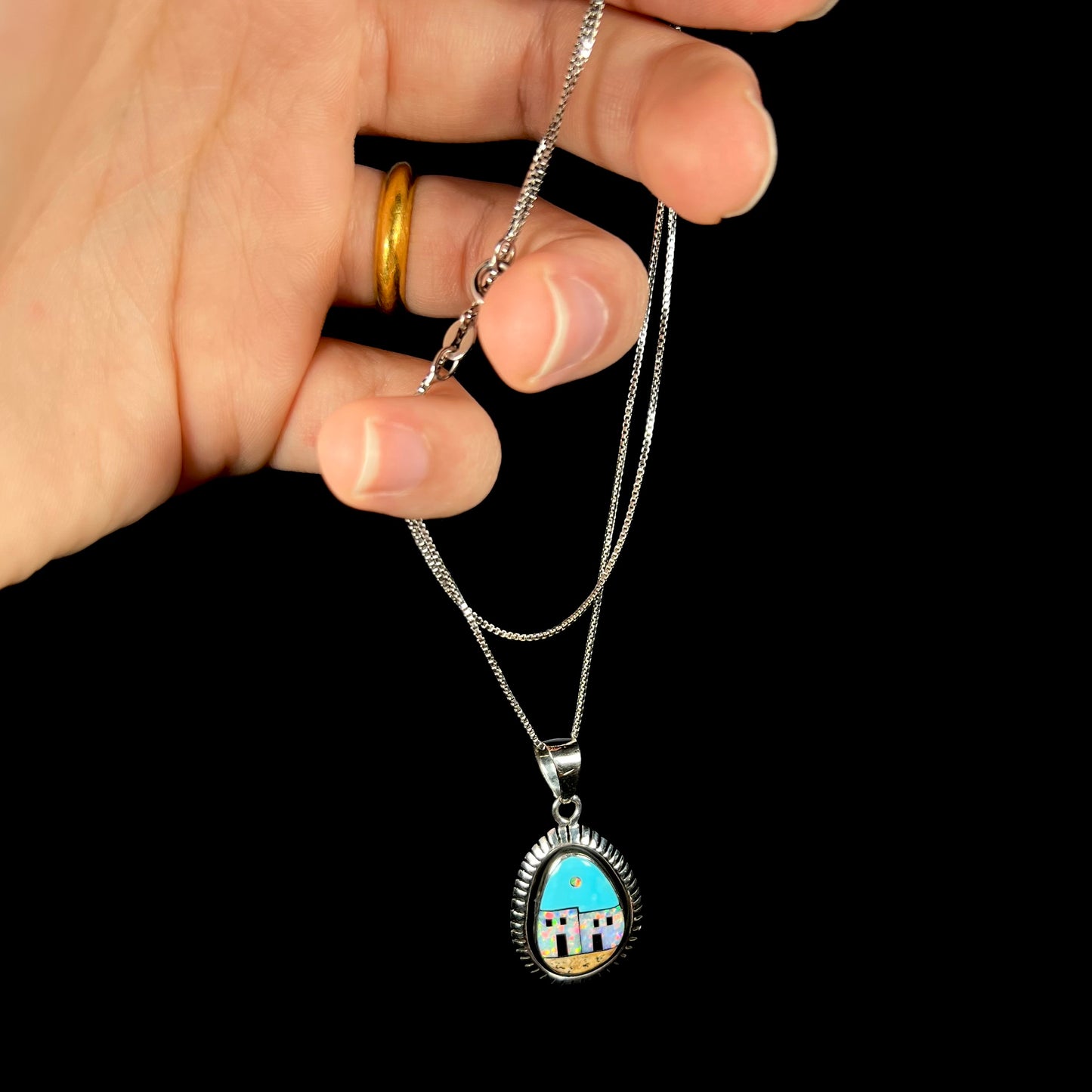A sterling silver Southwest-style opal and turquiose inlay pendant, held by a hand from its chain.