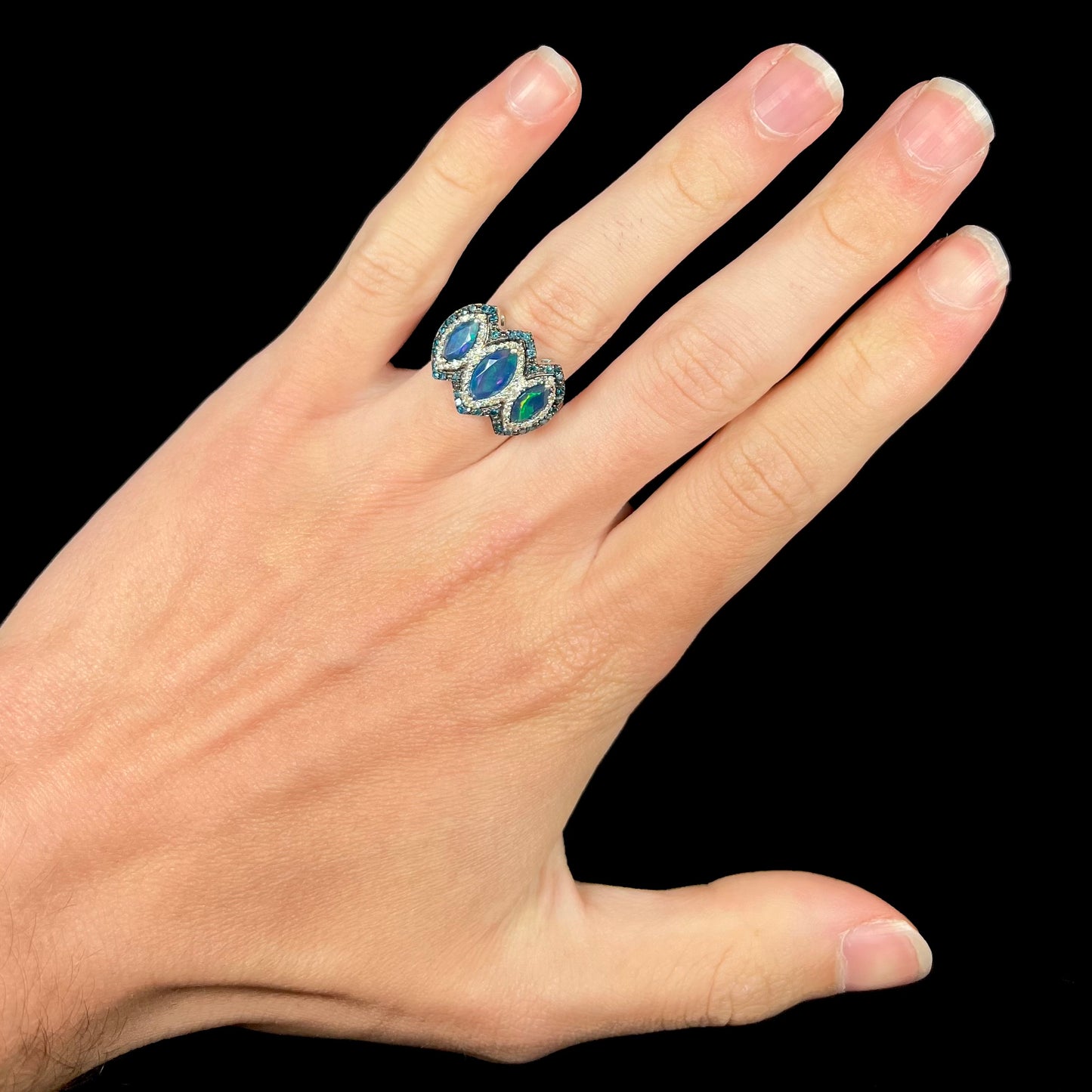 A three-stone blue opal ring with diamond accents worn on a hand against a black background.