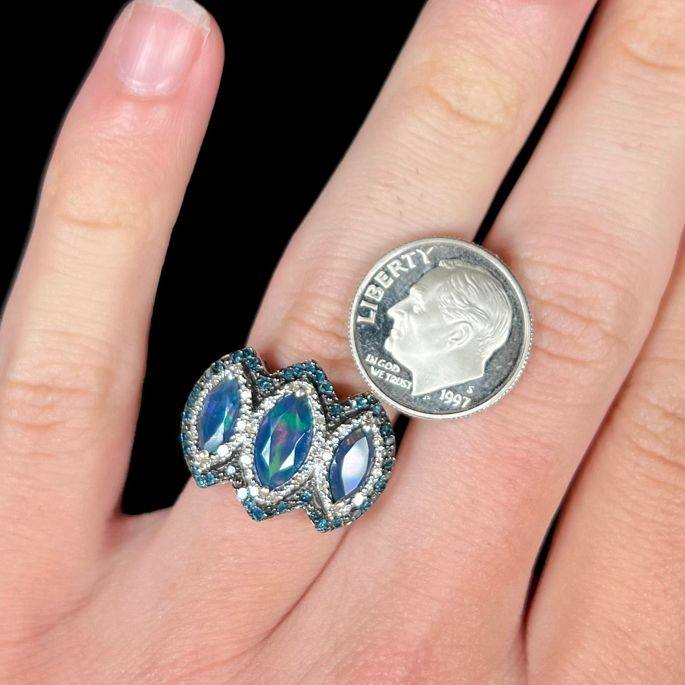 A three-stone blue opal and diamond ring worn on a hand next to a dime coin for scale.