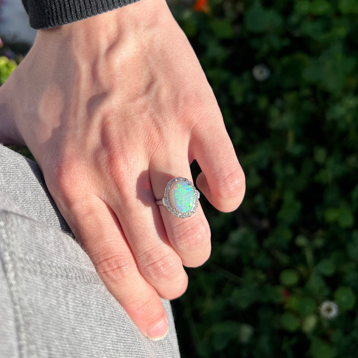 Hand resting on person's hip in sunlight wearing a vintage opal and diamond halo engagement ring with soft green play-of-color.