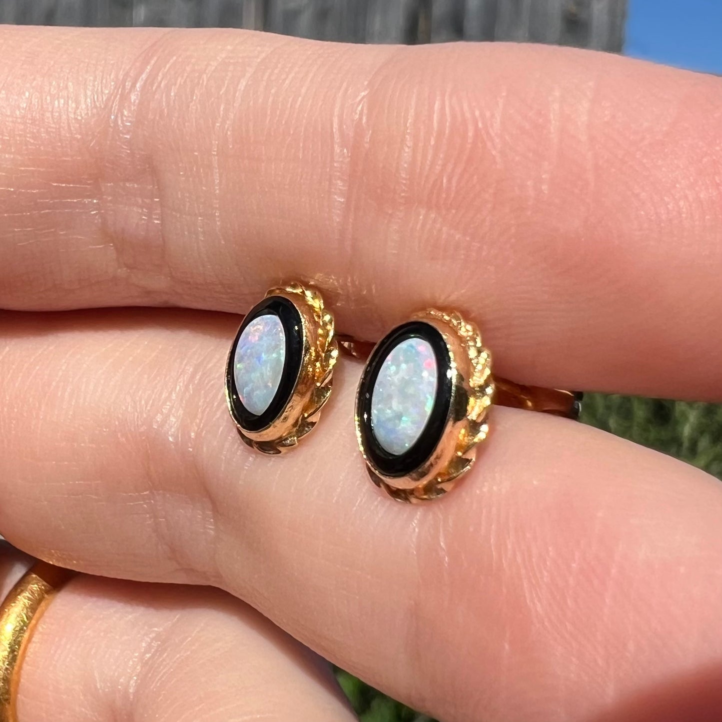 Angled close-up of vintage opal and onyx stud earrings held between two fingers in sunight.