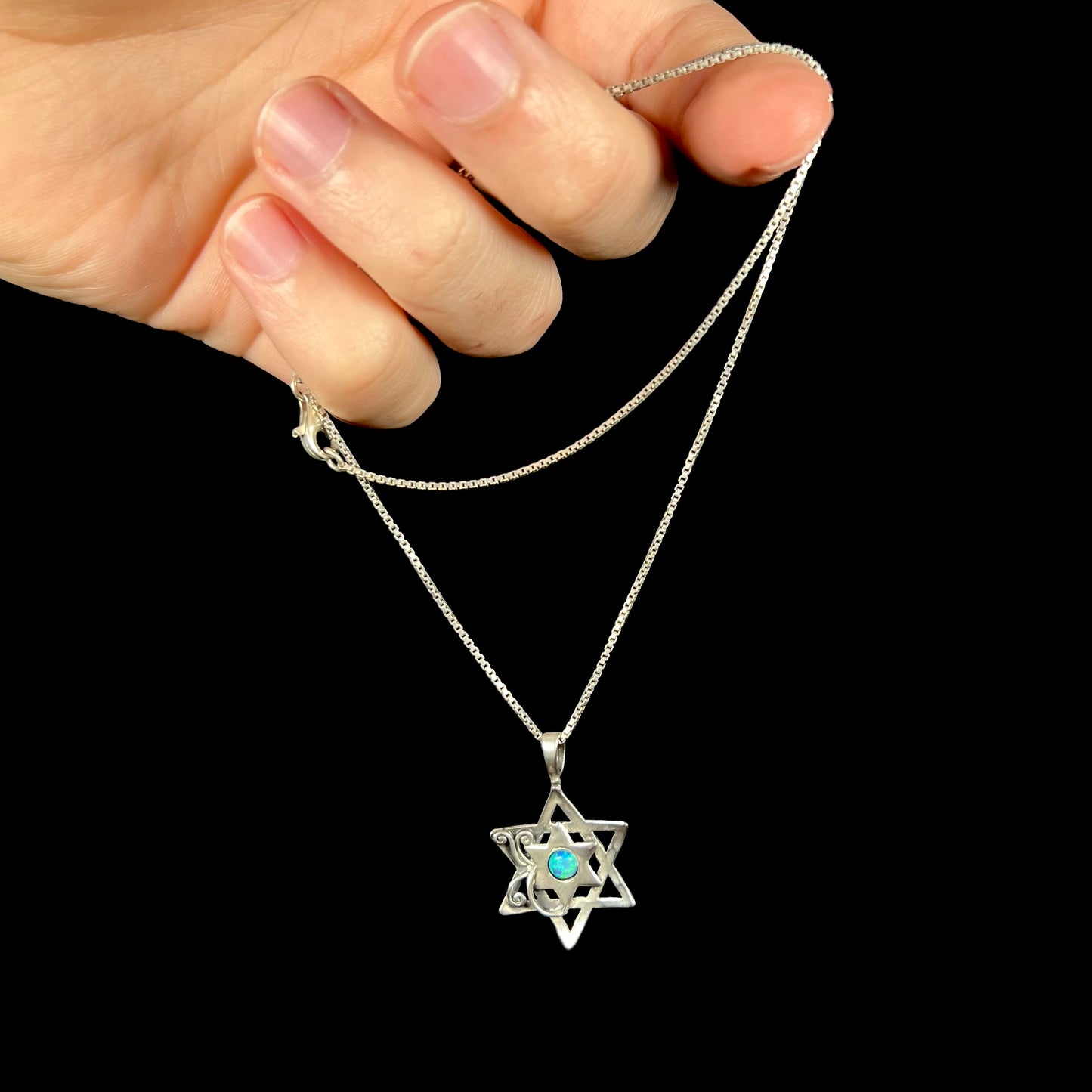 A vintage Star of David necklace set with a round blue lab-created opal, held in a hand by the chain.