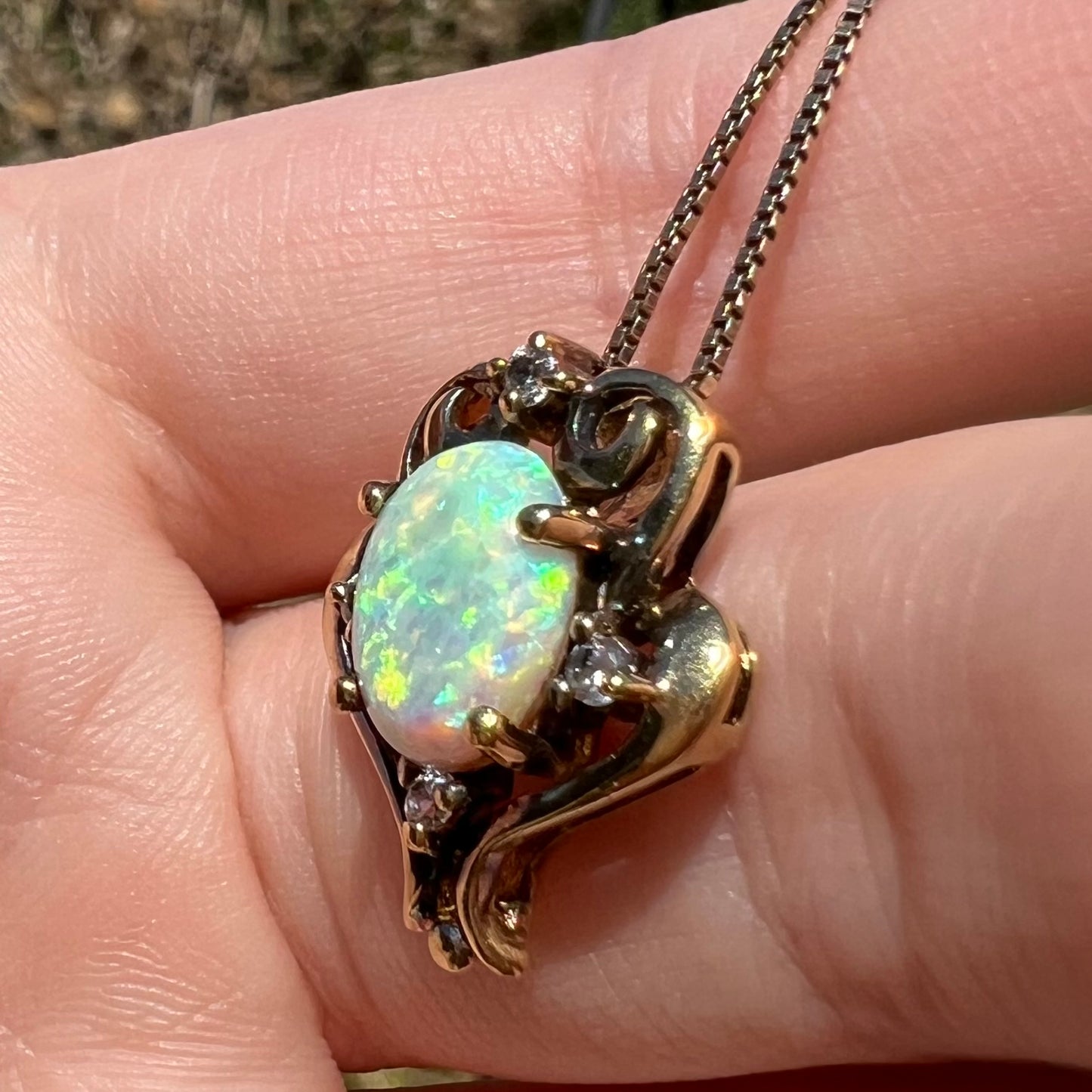 A vintage synthetic opal and CZ pendant with an aged patina over gold-tone metal, shown from an angle close-up in sunlight.
