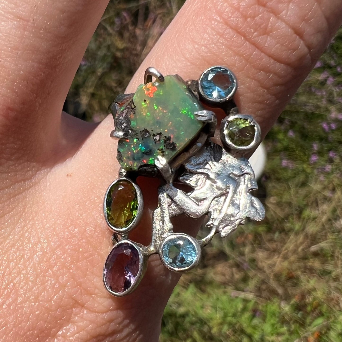 A silver men's ring set with rough Virgin Valley opal and gems with a female figure design shown close-up in sunlight.