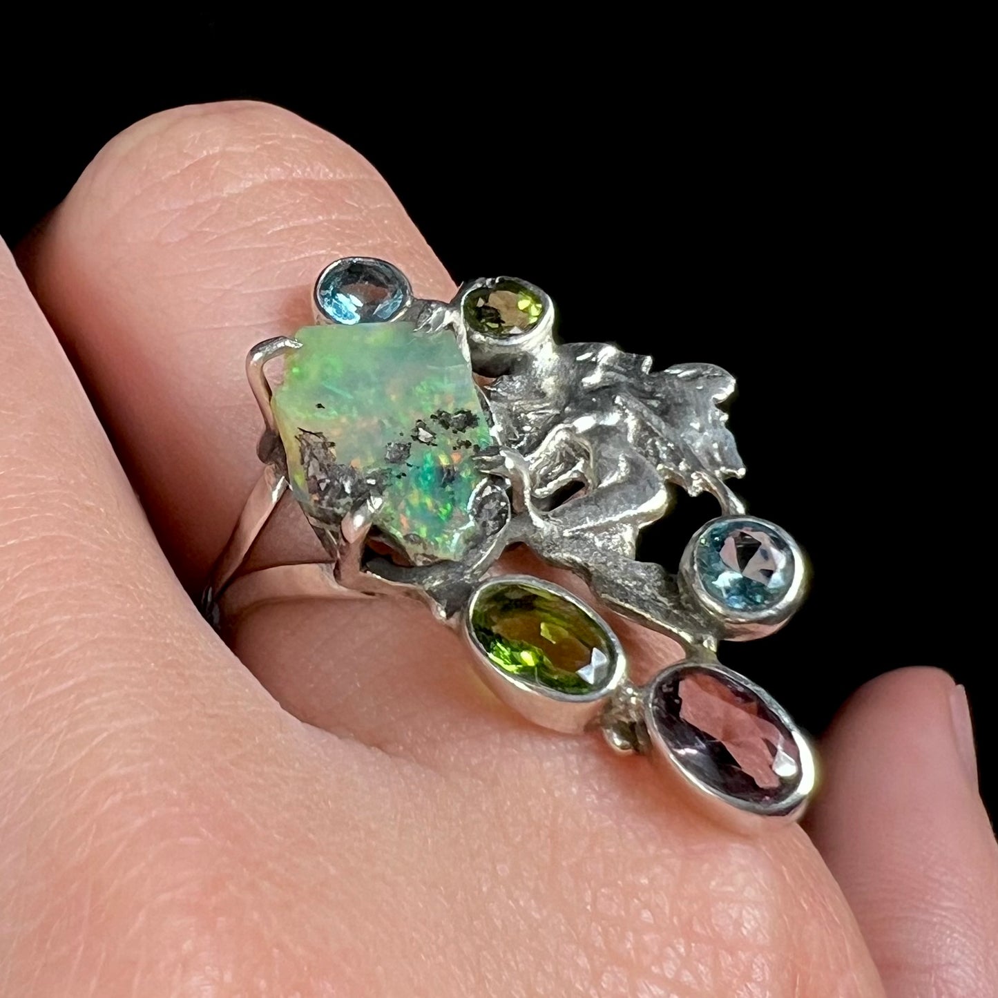 A silver men's ring set with rough Virgin Valley opal and gems with a female figure design worn on a hand at an angle.