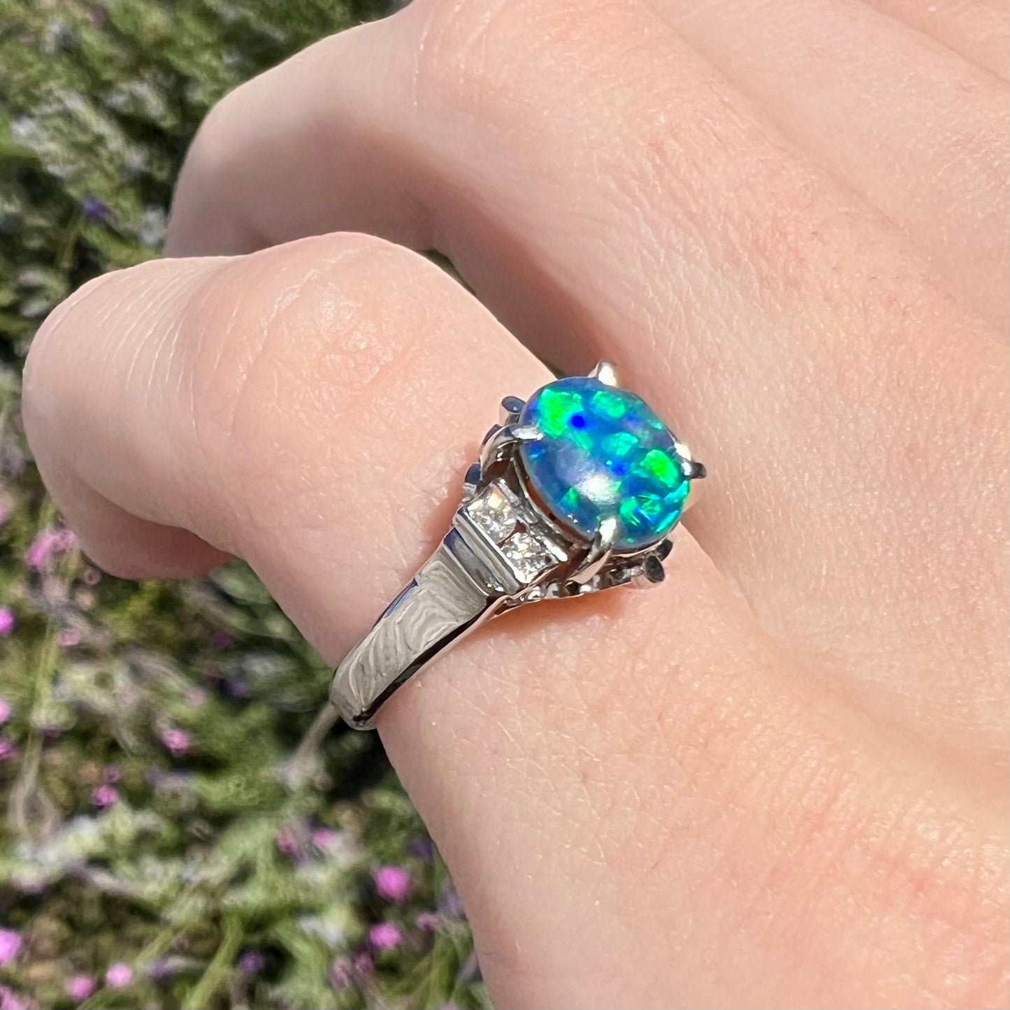 Vivid green and blue colors play across an oval-cut Lightning Ridge black opal, mounted in a platinum and diamond-accented ring worn on a pinkie.