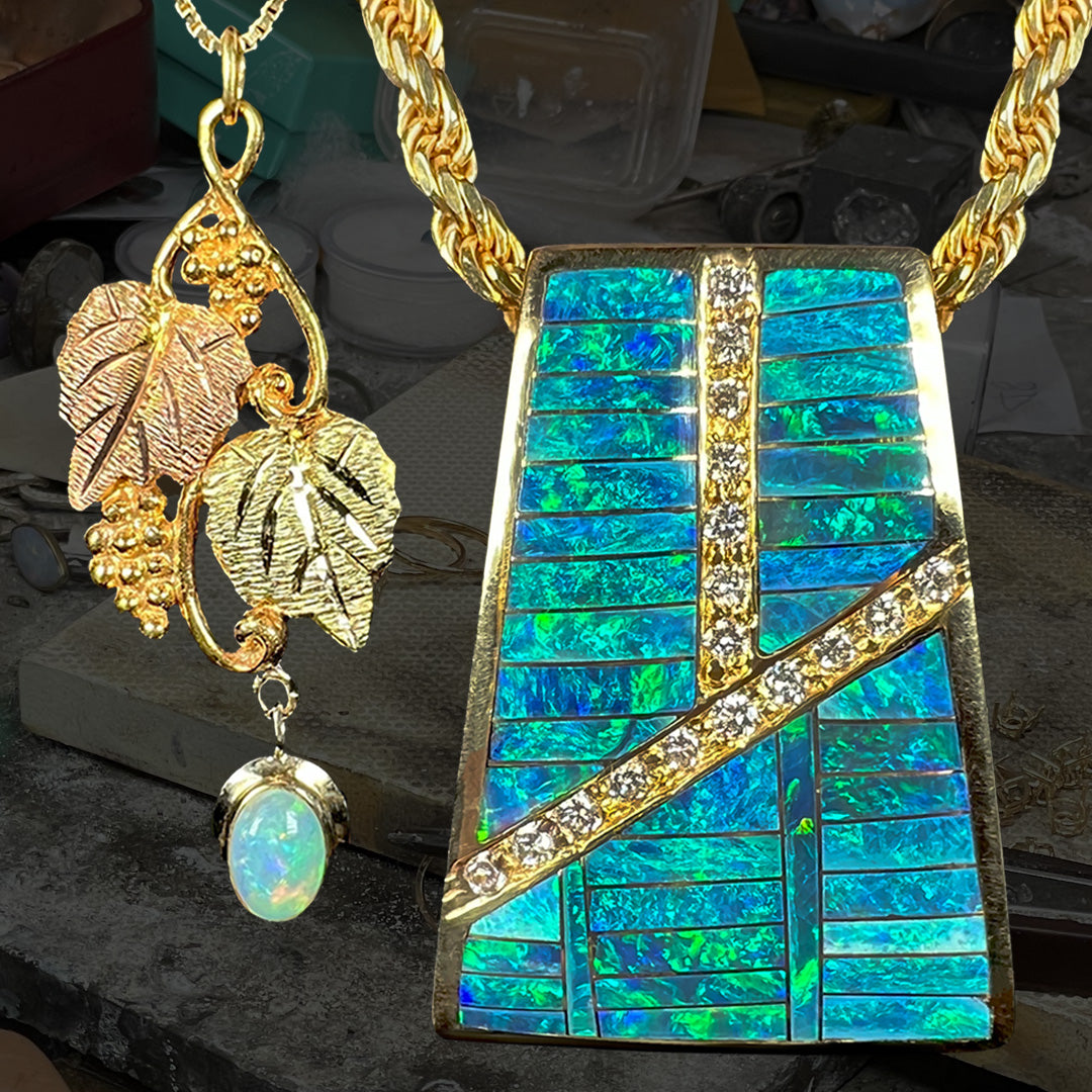 A Black Hills gold opal pendant shown to the left of a men’s gold Australian opal inlay and diamond necklace on a gold rope chain; both necklaces are in front of a darkened jeweler’s workbench.